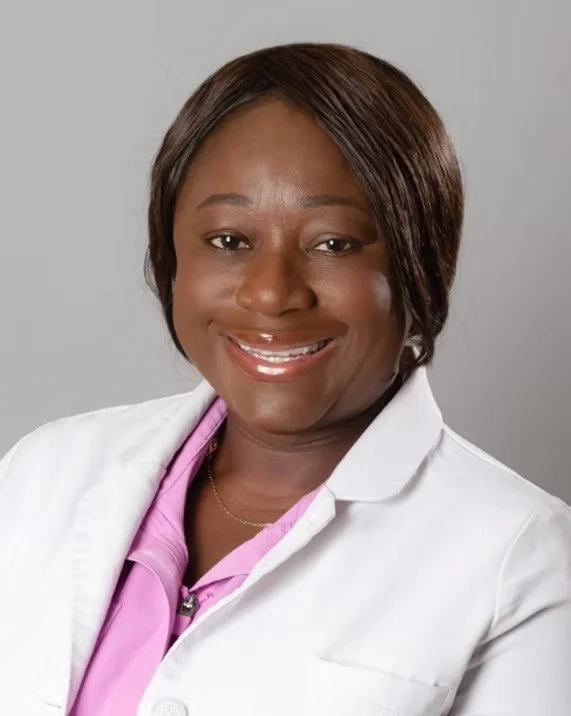 A woman with short, dark hair smiling, wearing a white coat over a pink shirt, against a plain gray background.