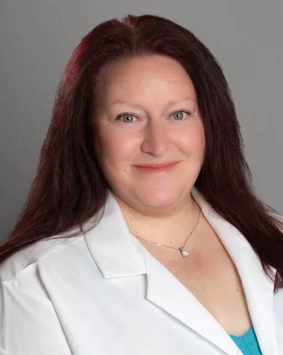 A woman with long dark red hair, blue eyes, and fair skin, wearing a white lab coat and a delicate necklace, smiling at the camera against a gray background.