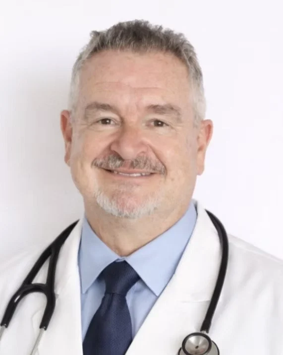 Smiling middle-aged male doctor with gray hair and beard, wearing a white lab coat, blue shirt, dark tie, and stethoscope around his neck, standing against a plain white background.