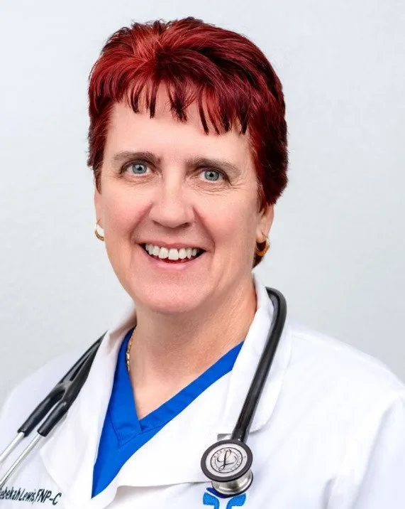 A woman with short red hair, wearing a white medical coat and blue scrubs, with a stethoscope around her neck, smiling at the camera.