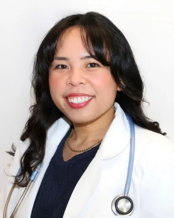 A smiling female doctor with long black hair wearing a white lab coat and a stethoscope around her neck, against a plain white background.