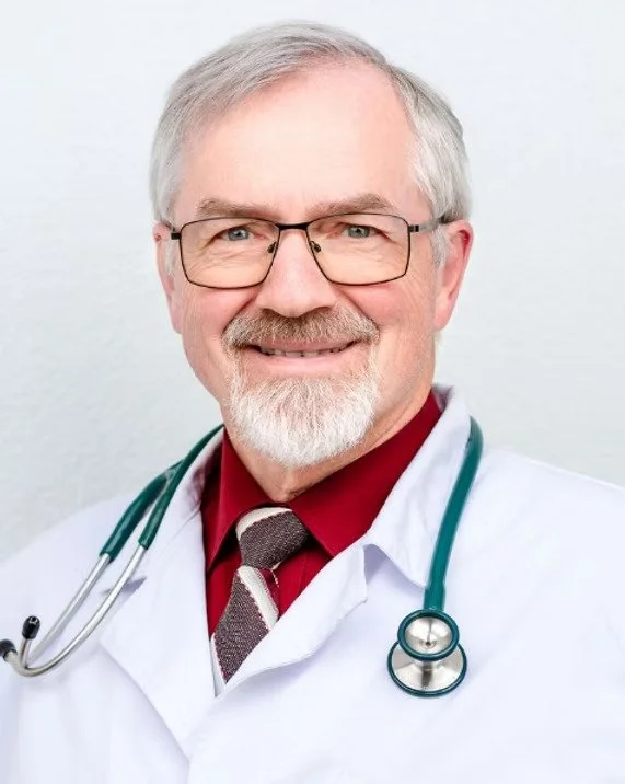 A smiling male doctor with glasses, gray hair, and a beard, wearing a white coat and a stethoscope around his neck.