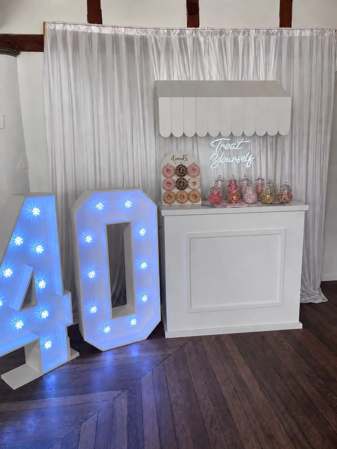 Decorative display with large illuminated numbers '40', a white sweet stall with glass jars filled with sweets, plus a sign that reads 'donuts' with donuts on, and a neon sign that says 'Treat Yourself,'