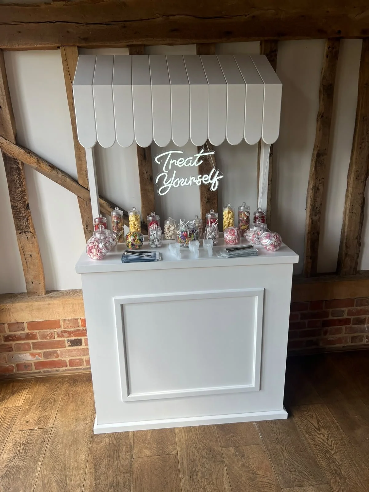 A white treat stand with a scalloped canopy and a neon sign that says 'Treat Yourself'. It displays jars of candies, including pink and white cotton candy, gummies, and colorful candies, along with small serving utensils and napkins.