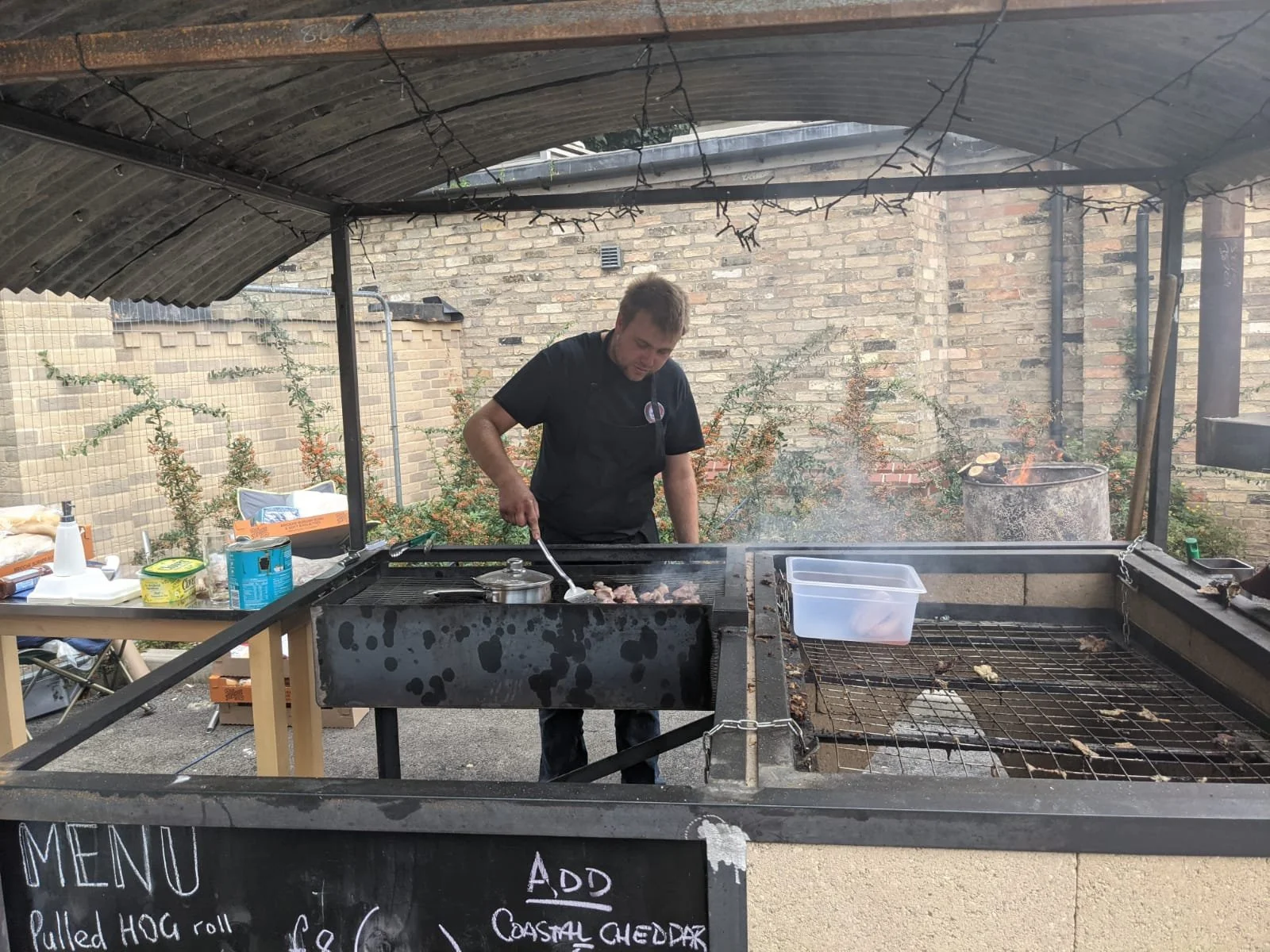 Man cooking meat on outdoor grill under a shaded canopy with a brick wall in the background.