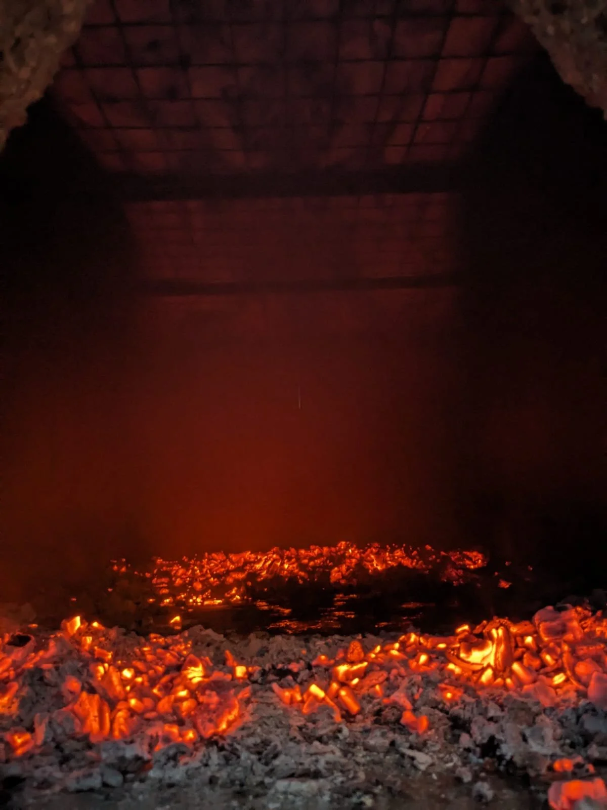 Hot coals in a fire with glowing embers and ashes under a roof structure, illuminated orange from the fire.
