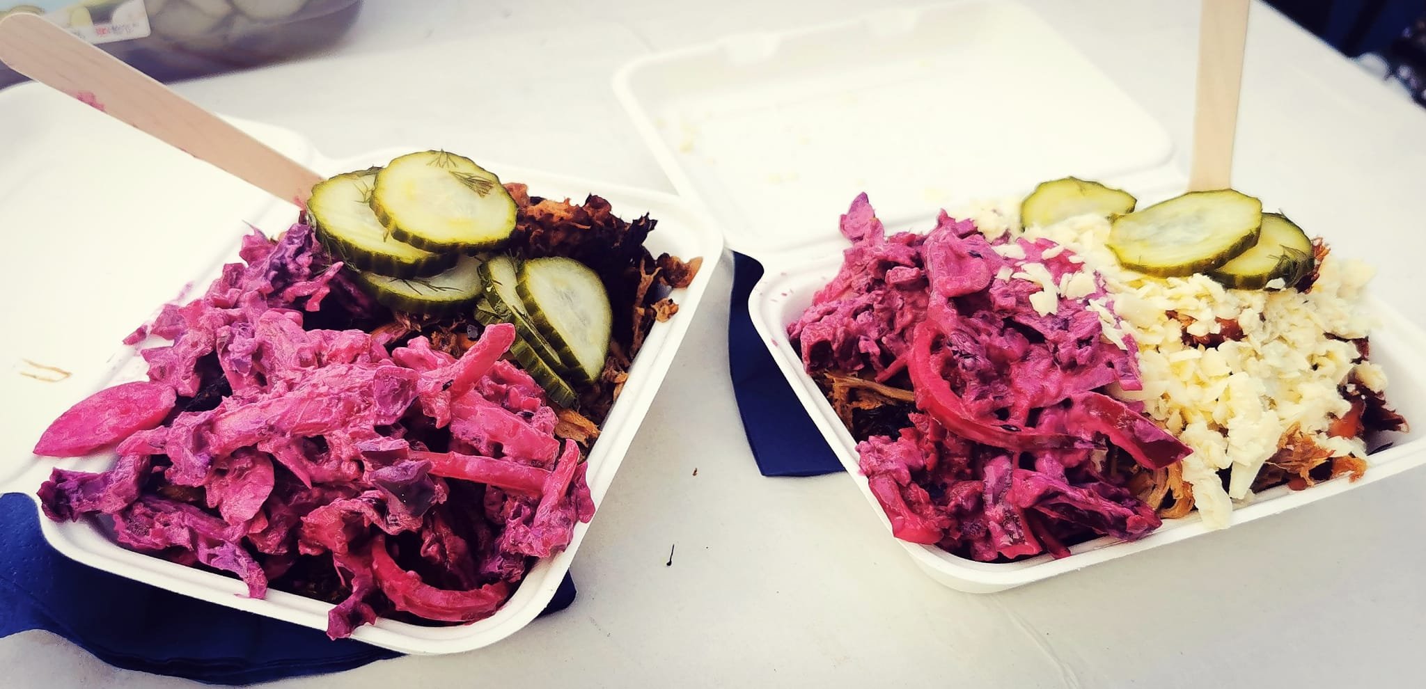 Two takeout containers filled with shredded pink-colored coleslaw, topped with pickle slices, placed on a white surface.