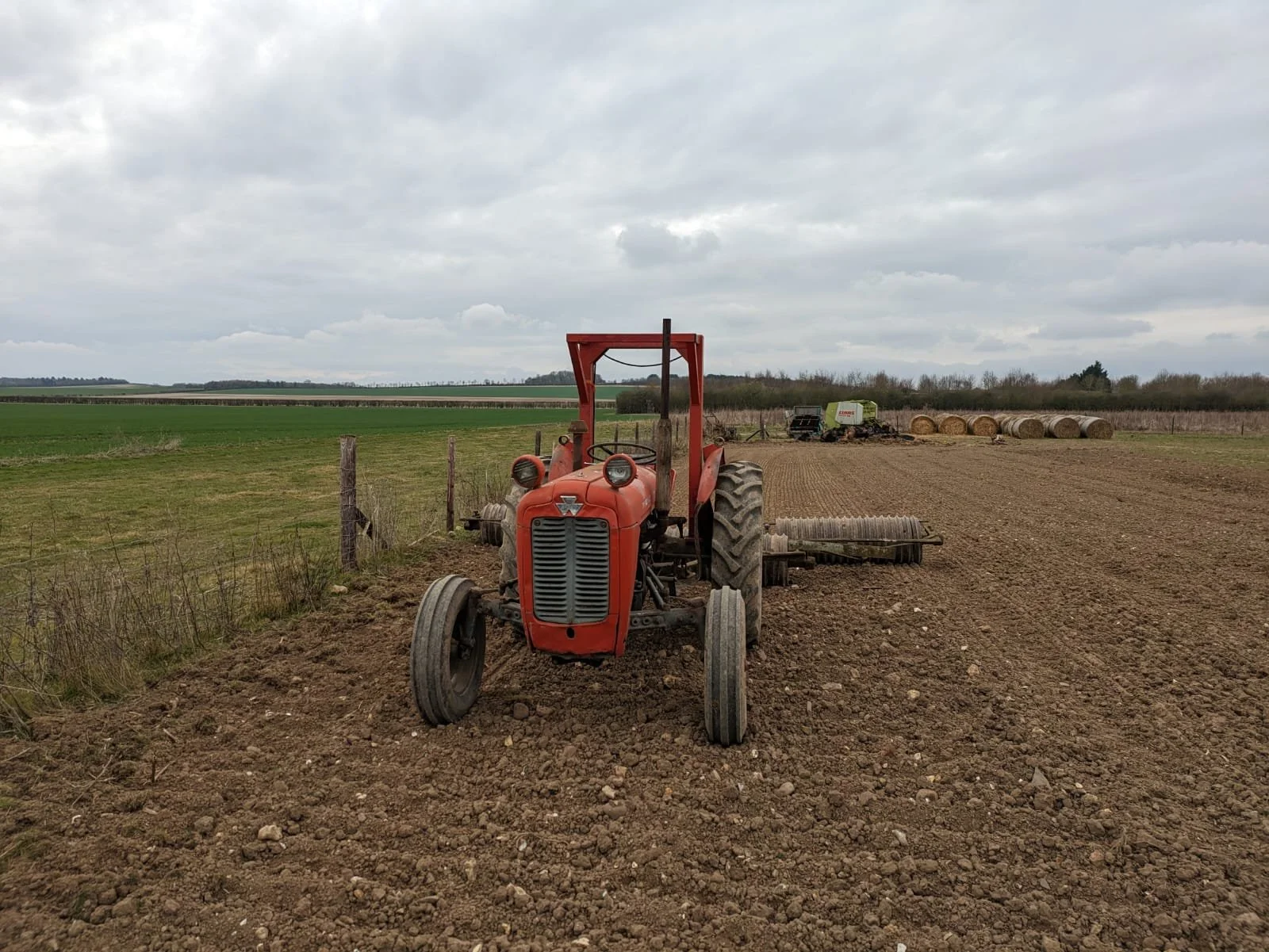 Red tractor on a farm field under a cloudy sky, with hay bales in the distance.