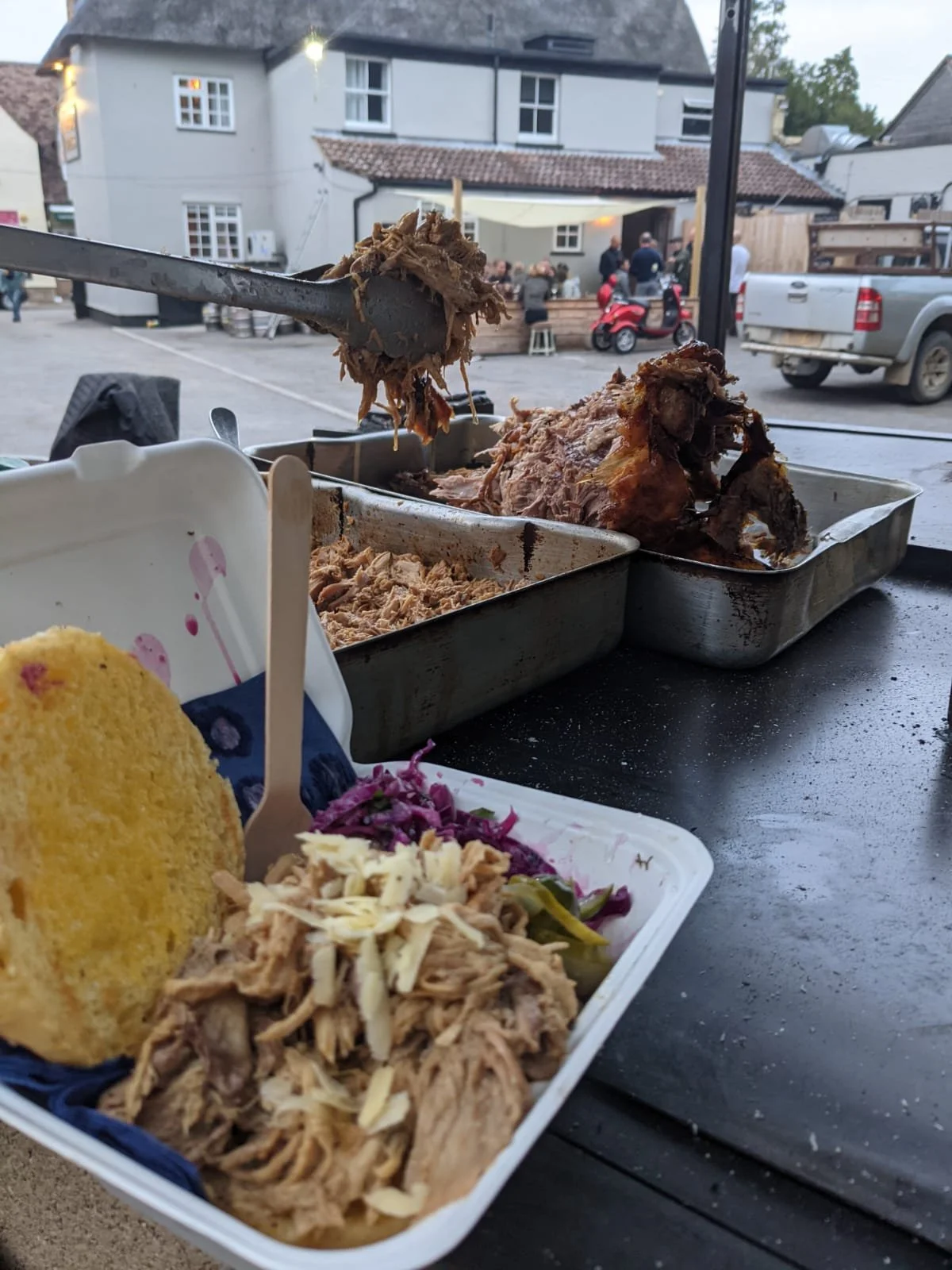 Tray of shredded barbecue pork with coleslaw and cornbread in a white container, and a metal tray of pulled pork in the background, with a street scene outside.