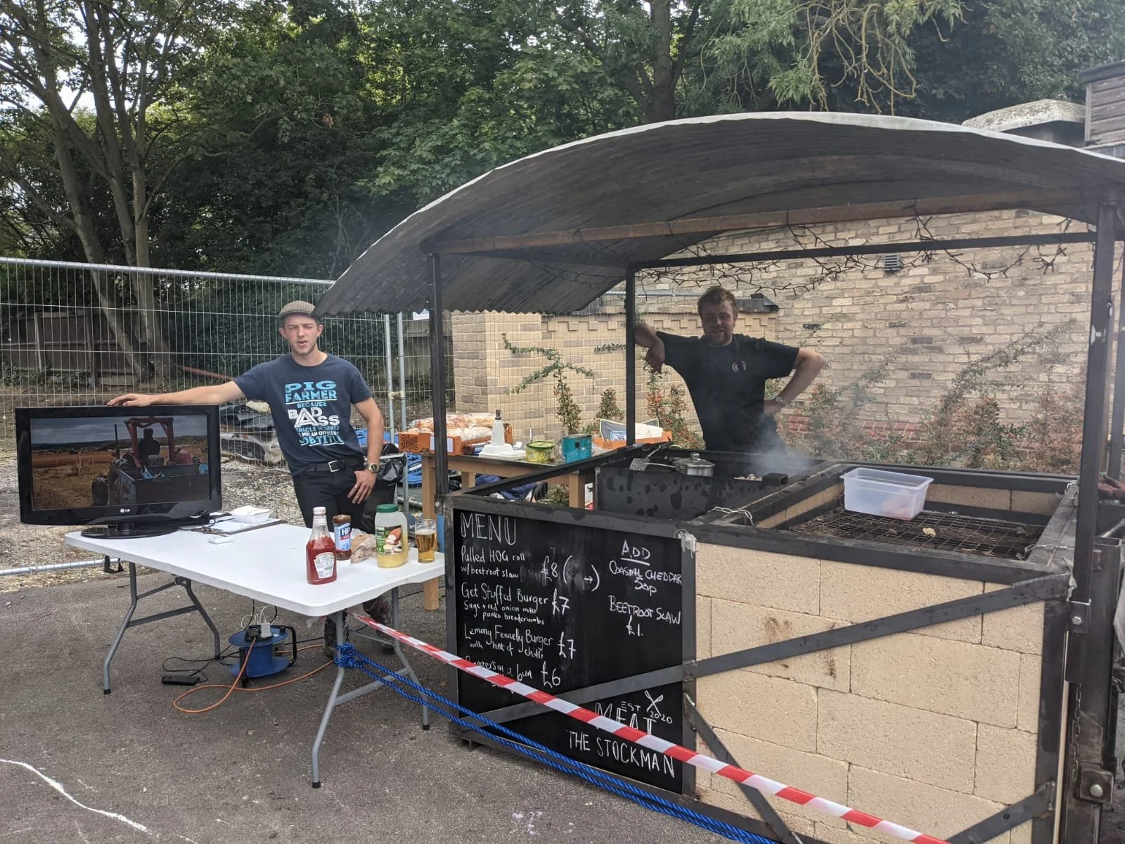 Two men stand behind a barbecue stand, one on each side. The man on the left is wearing a gray cap and a blue t-shirt, and the man on the right is cooking on a grill under a canopy. There is a small television on the left side and various condiments on a white table in front of it. The scene appears to be at an outdoor event.