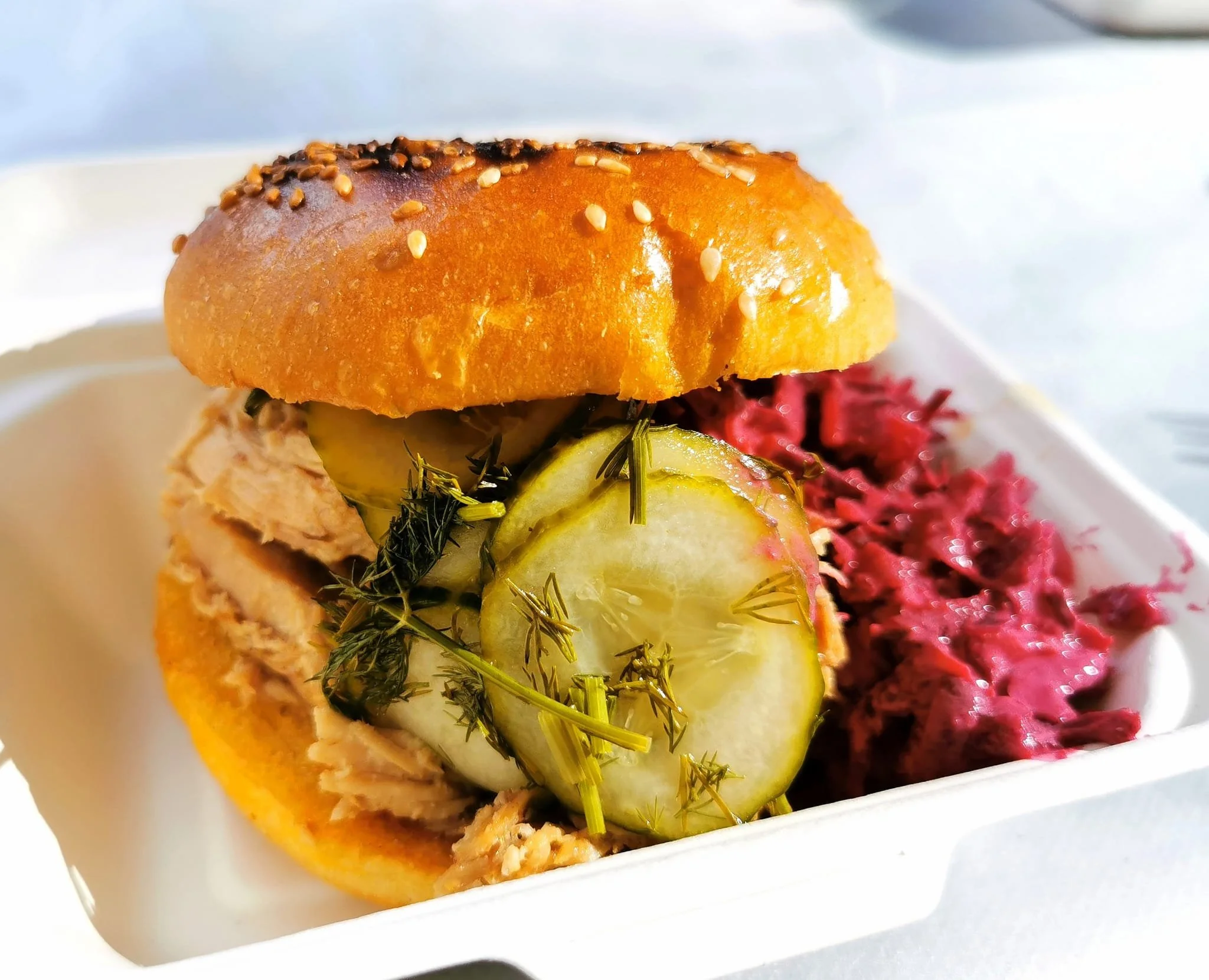 Sandwich with pickles, herbs, and shredded meat, served with a side of beet salad in a white container.