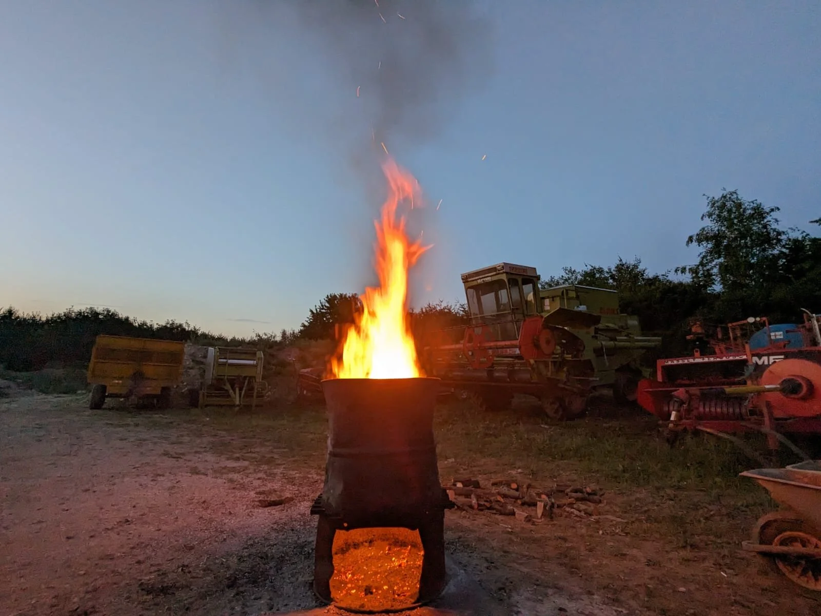 A fire burning inside a metal drum with sparks flying upward, surrounded by vintage farming equipment and machinery outdoors at dusk.