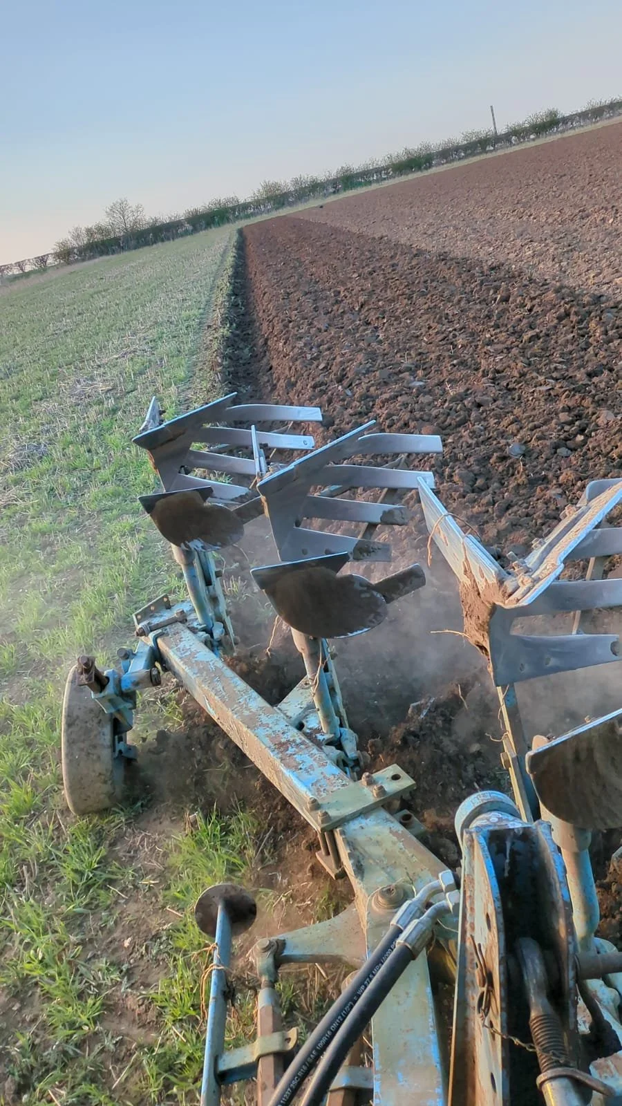 A tractor plowing a field with freshly tilled dark soil on the right and green grass on the left, under a clear blue sky.