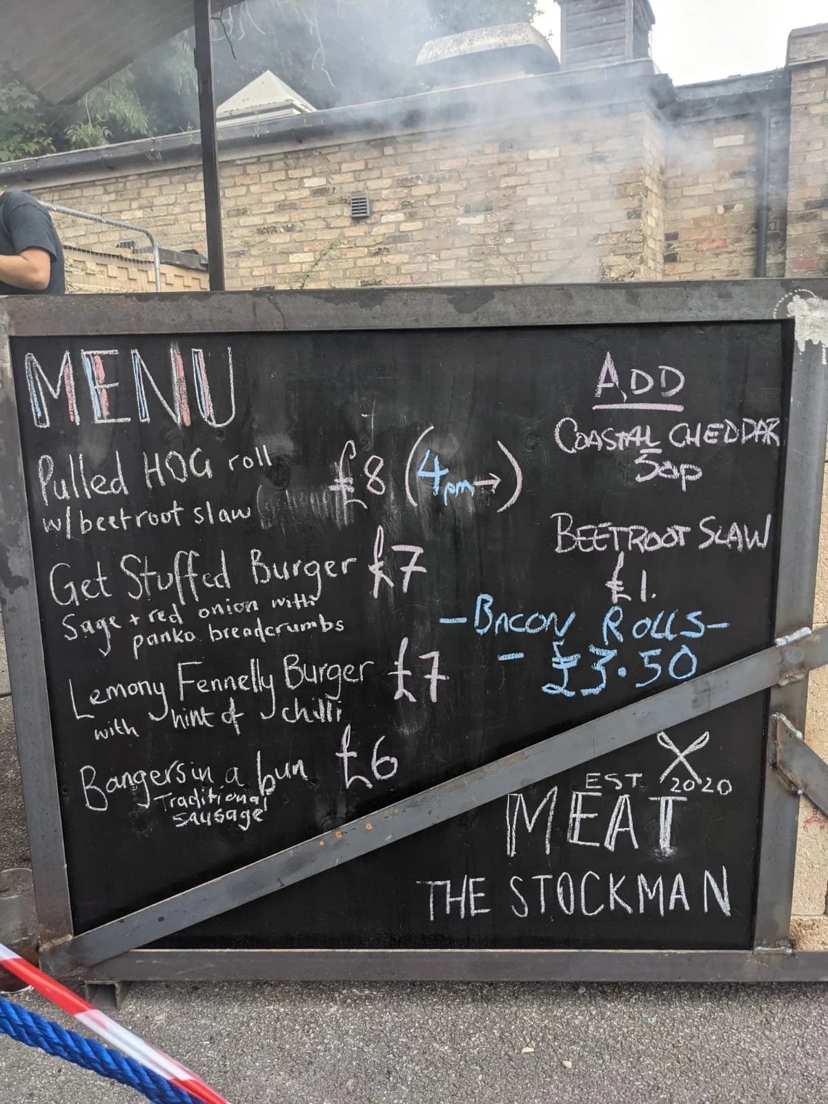 Black chalkboard menu with colorful handwritten items, listing various burger options and prices, including an add-on section for coastal cheddar soup, beetroot slaw, bacon rolls, and a note about traditional sausage.