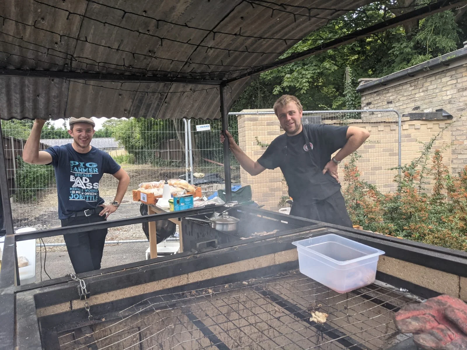 Two men standing behind a barbecue grill at an outdoor event, smiling. One man is wearing a blue t-shirt with a graphic print, the other is dressed in black with a black apron. There are food items and condiments on a table behind them, and a metal fence in the background.