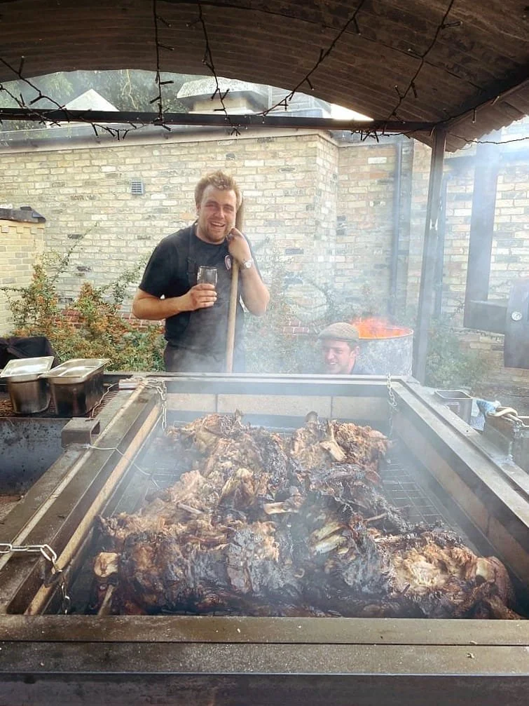 Two men smiling in a backyard barbecue area with a large grill loaded with cooked meat, one holding a glass, one with a tool, with smoke and a red fire in the background.