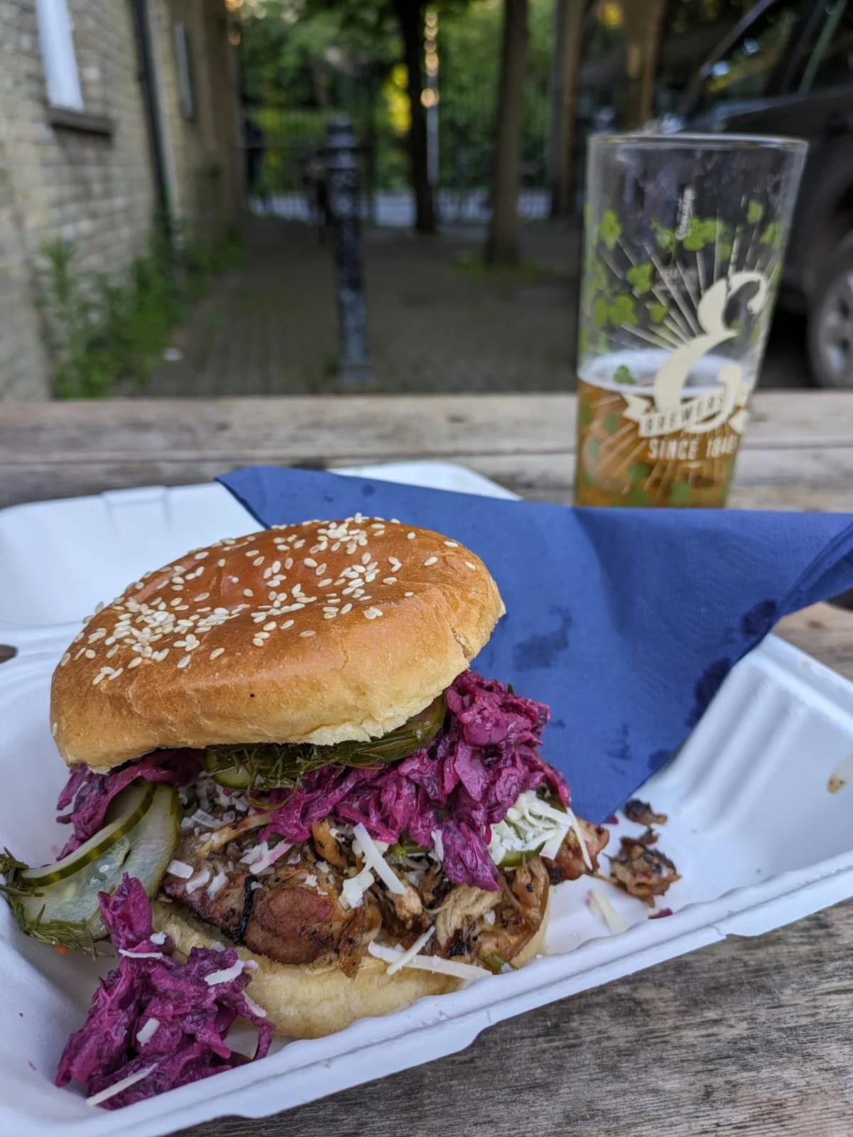 A pulled pork sandwich with coleslaw and pickles in a burger bun with sesame seeds, served in a white foam tray on a wooden table, with a glass of beer in the background.