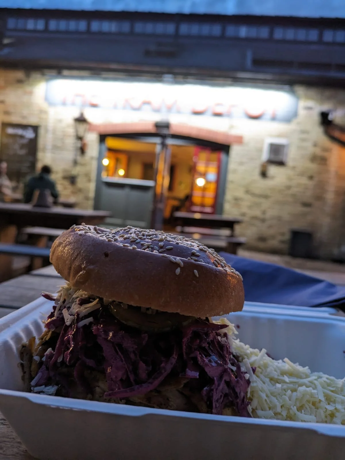 A sandwich with shredded meat, coleslaw, and pickles inside a sesame seed bun, served with shredded cheese on the side, on a white tray outside at a restaurant, with a blurred brick building and neon sign in the background.