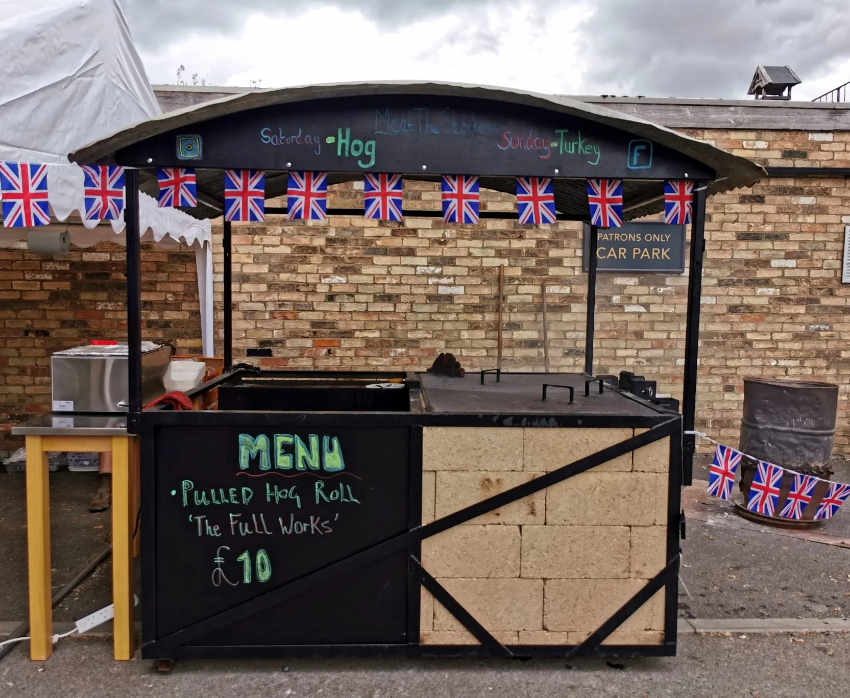 A black outdoor food stall decorated with British flags, serving pulled hog rolls for £10, with a menu listing the dish and its price, situated against a brick wall.
