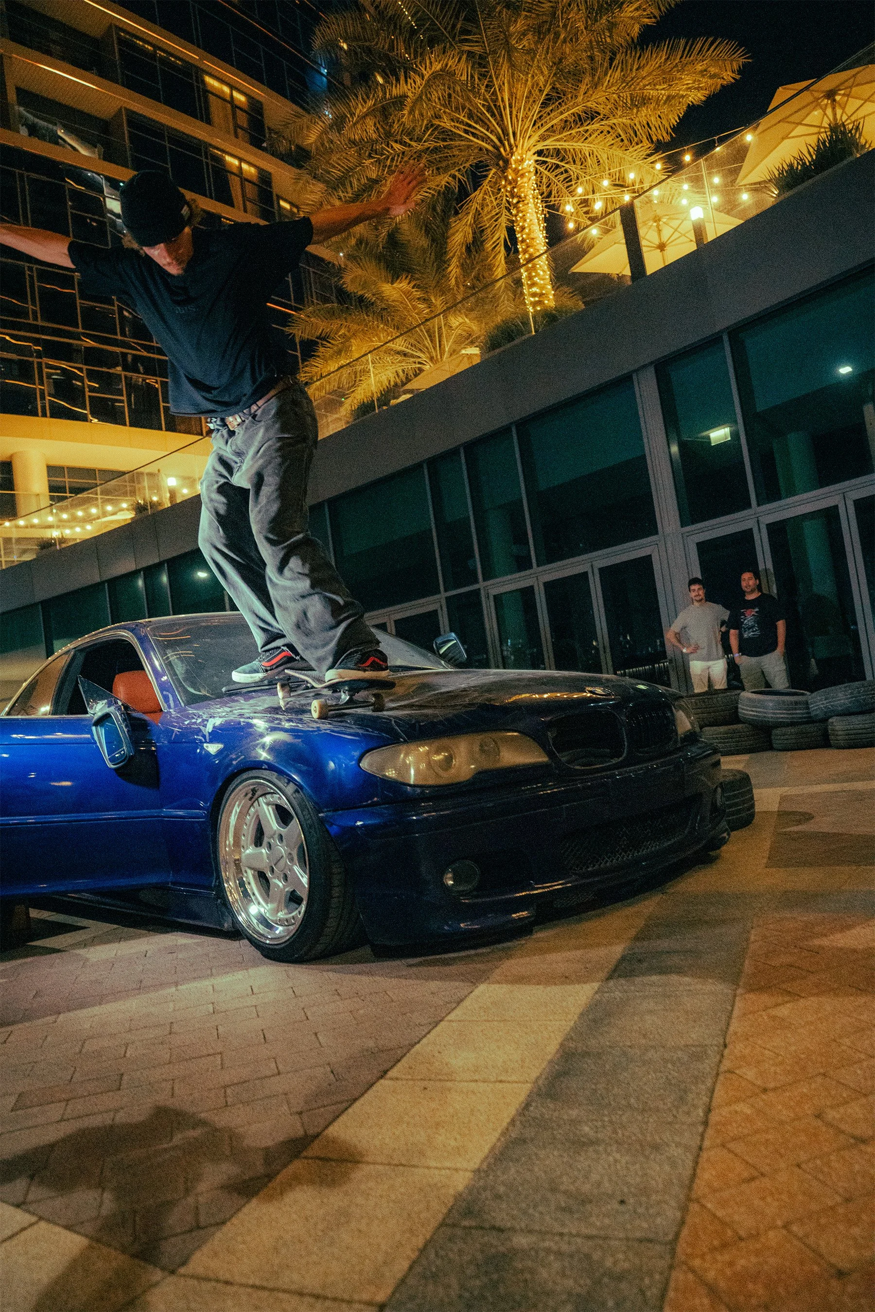 A person skating on the roof of a damaged black car amidst an urban night setting with palm trees, string lights, and two people observing in the background.
