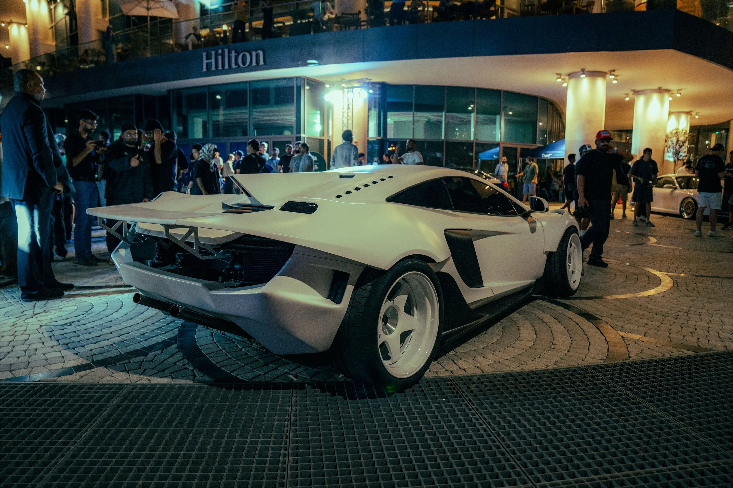 A white sports car with an aerodynamic design and a large rear wing is parked outside the Hilton hotel at night. The scene is lively with a crowd of people gathered around, some taking photos or videos. The hotel entrance is illuminated, and there are multiple floors with guests visible inside.