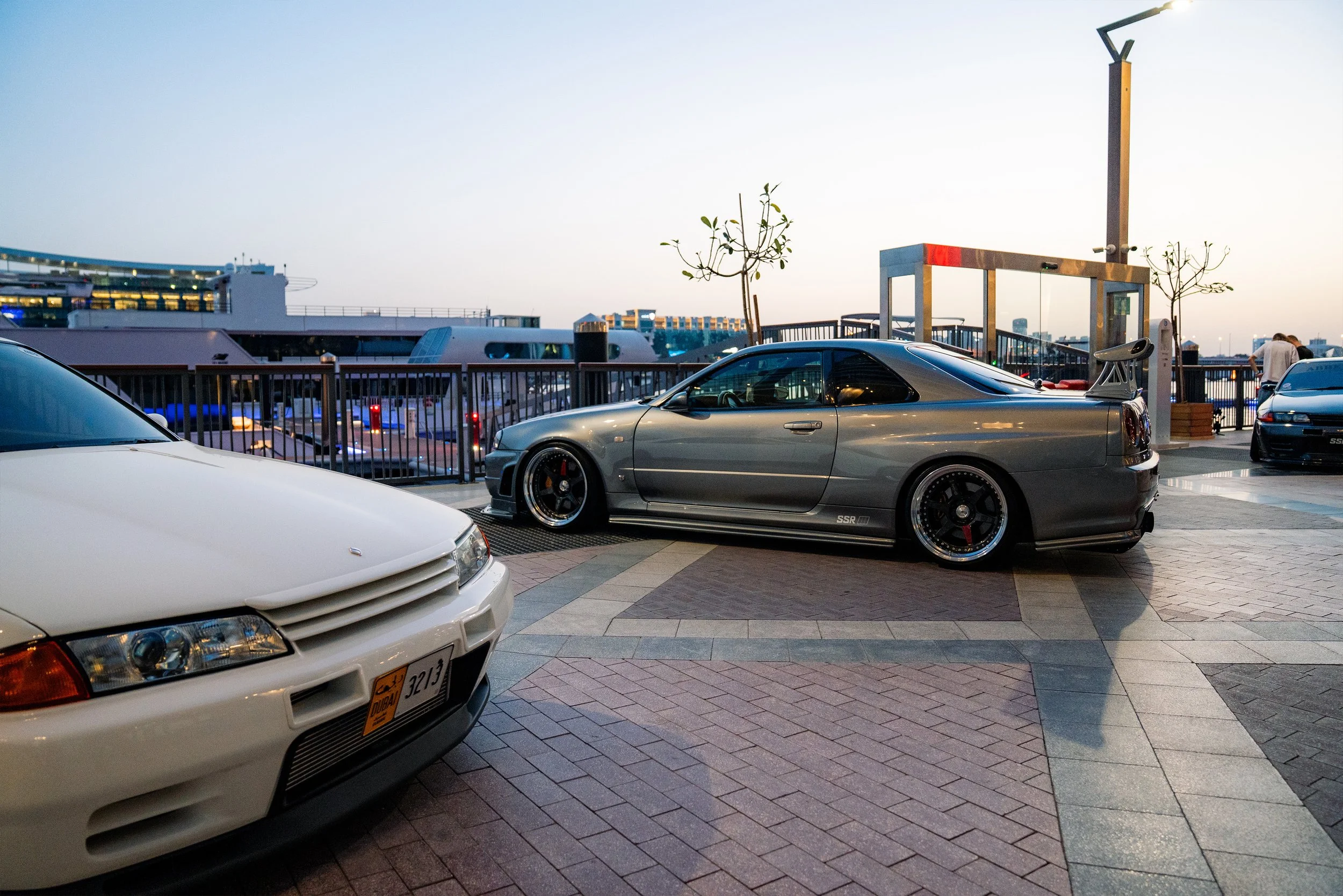 A parking lot on a boat deck with several cars, including a gray sports car with a spoiler, and a white car in the foreground. In the background, there are boats docked at the marina, a railing, and a clear sky at dusk.