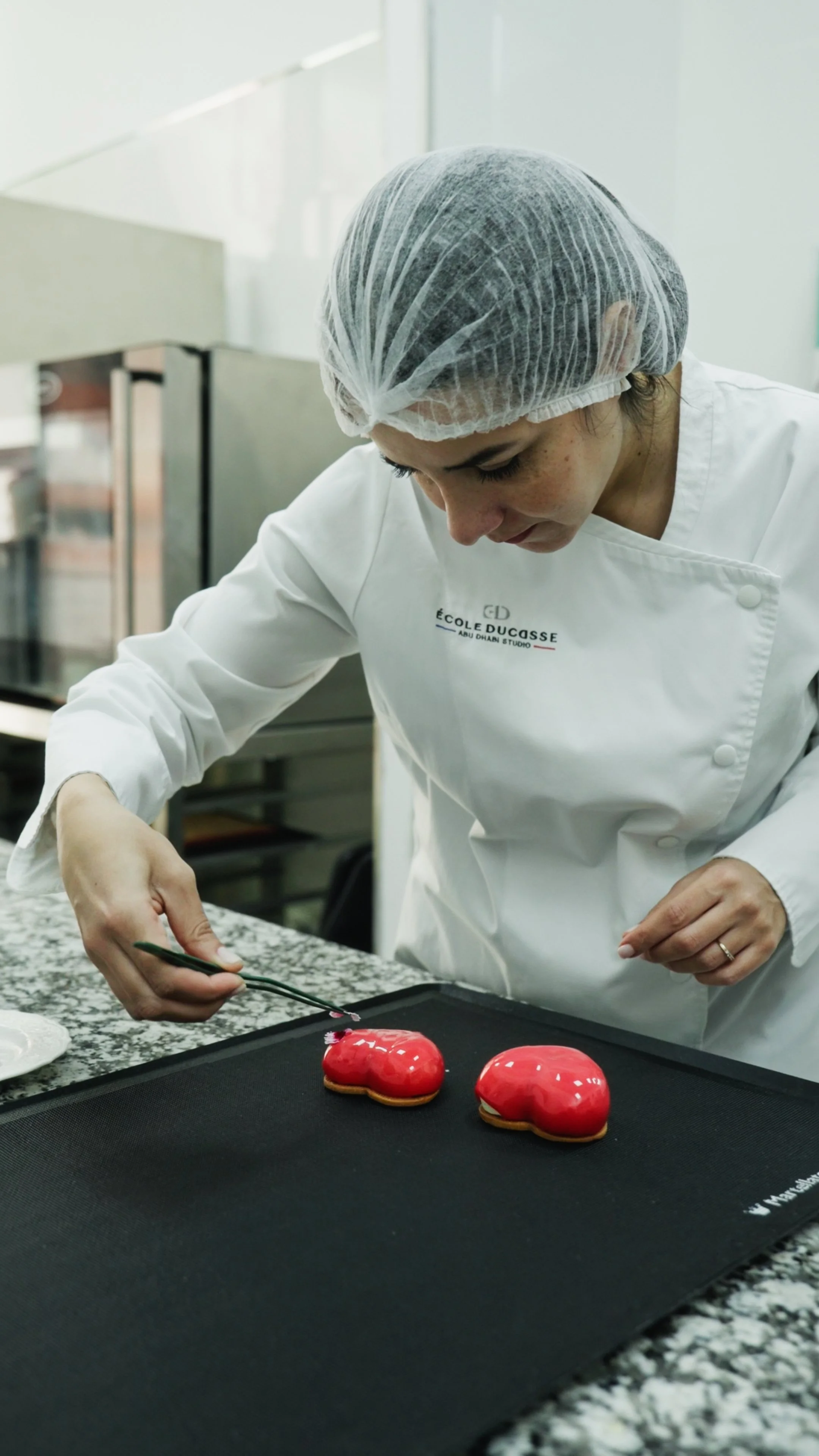 A baker in a white uniform and hairnet is decorating heart-shaped cookies with red glaze on a black tray.