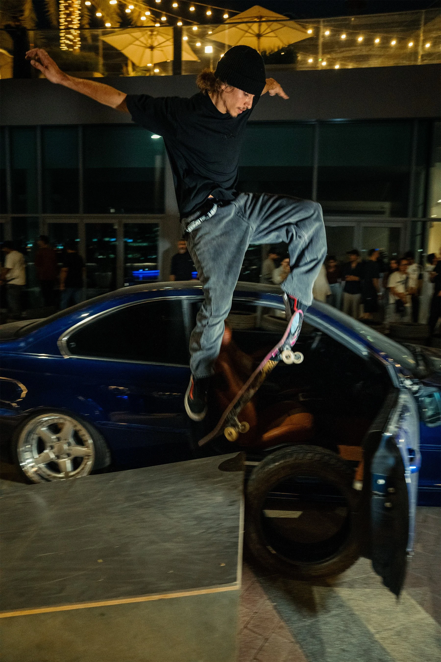 A young man skateboarding on top of a black sports car in an indoor space with glass windows and string lights.