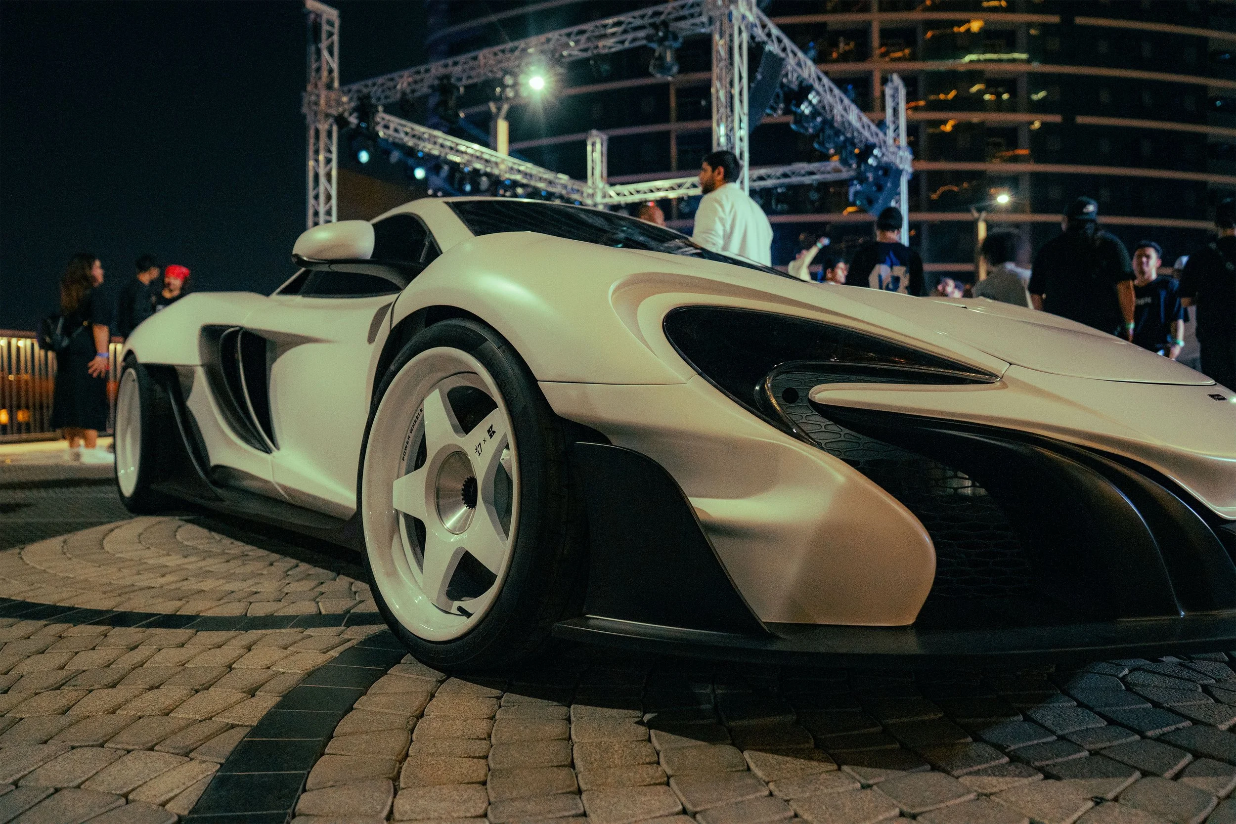 A white futuristic sports car displayed at night at an outdoor event with a stage and people in the background.