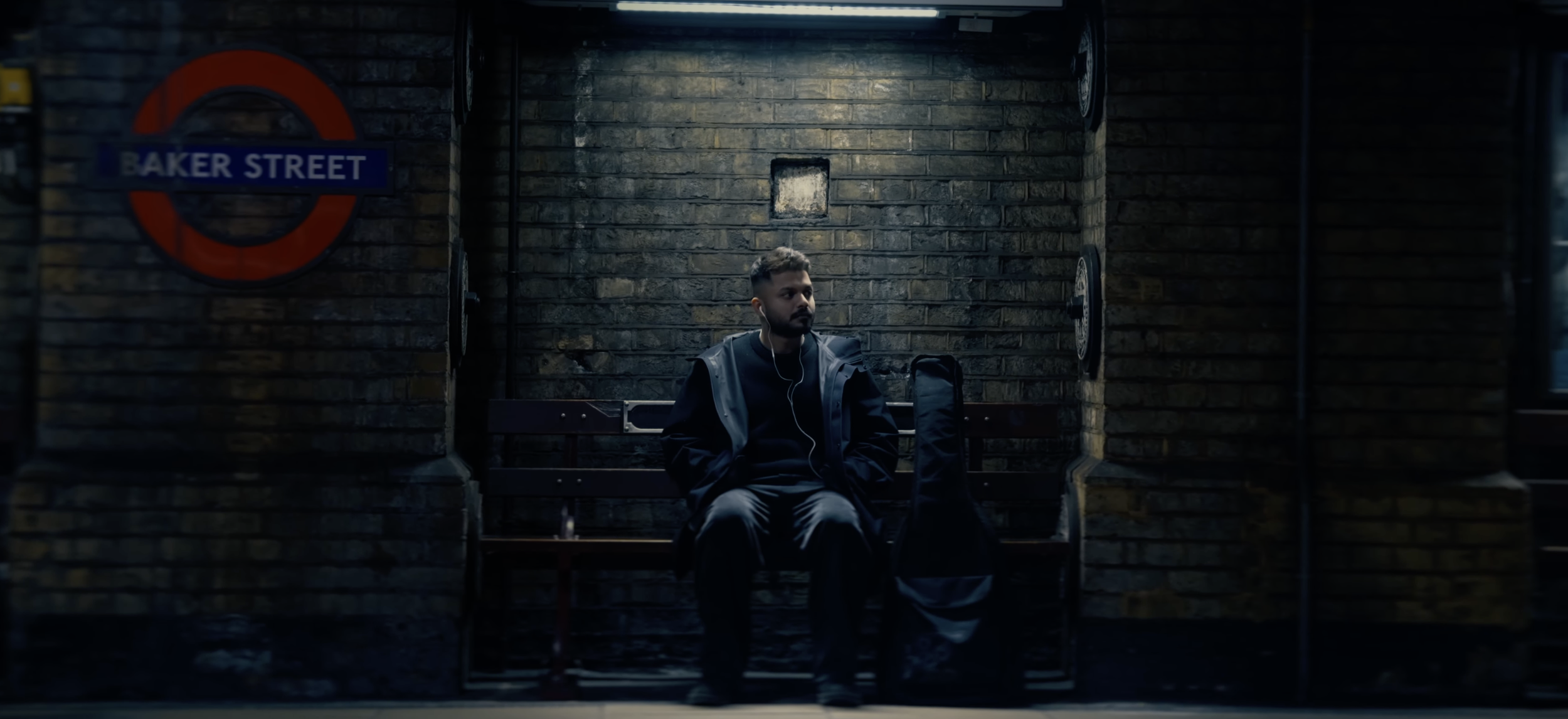 A young man sitting on a bench at a dimly lit underground subway station, wearing headphones and carrying a bag.