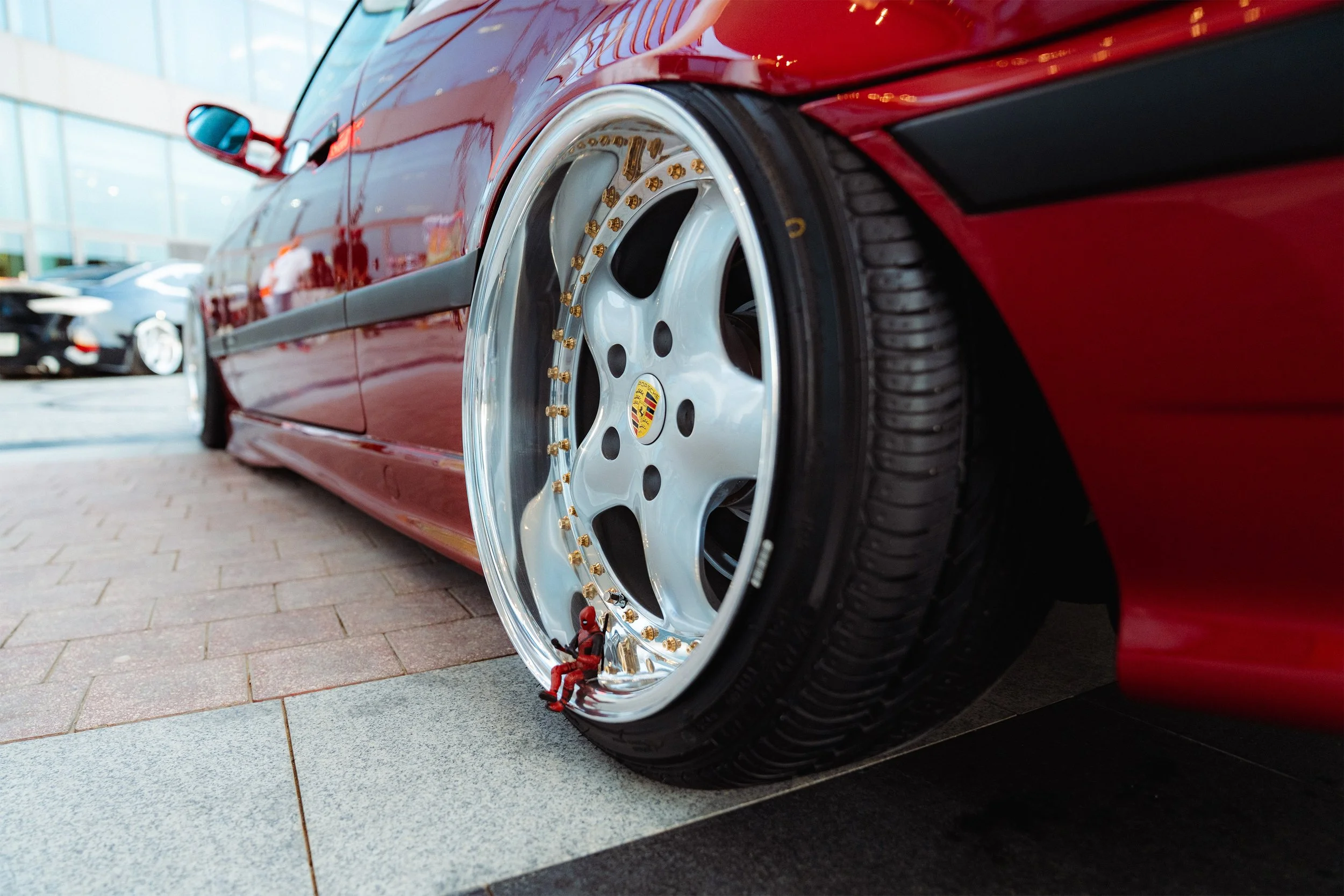 A close-up side view of a red sports car with custom wheels and gold lug nuts parked on a sidewalk in front of a modern glass building.
