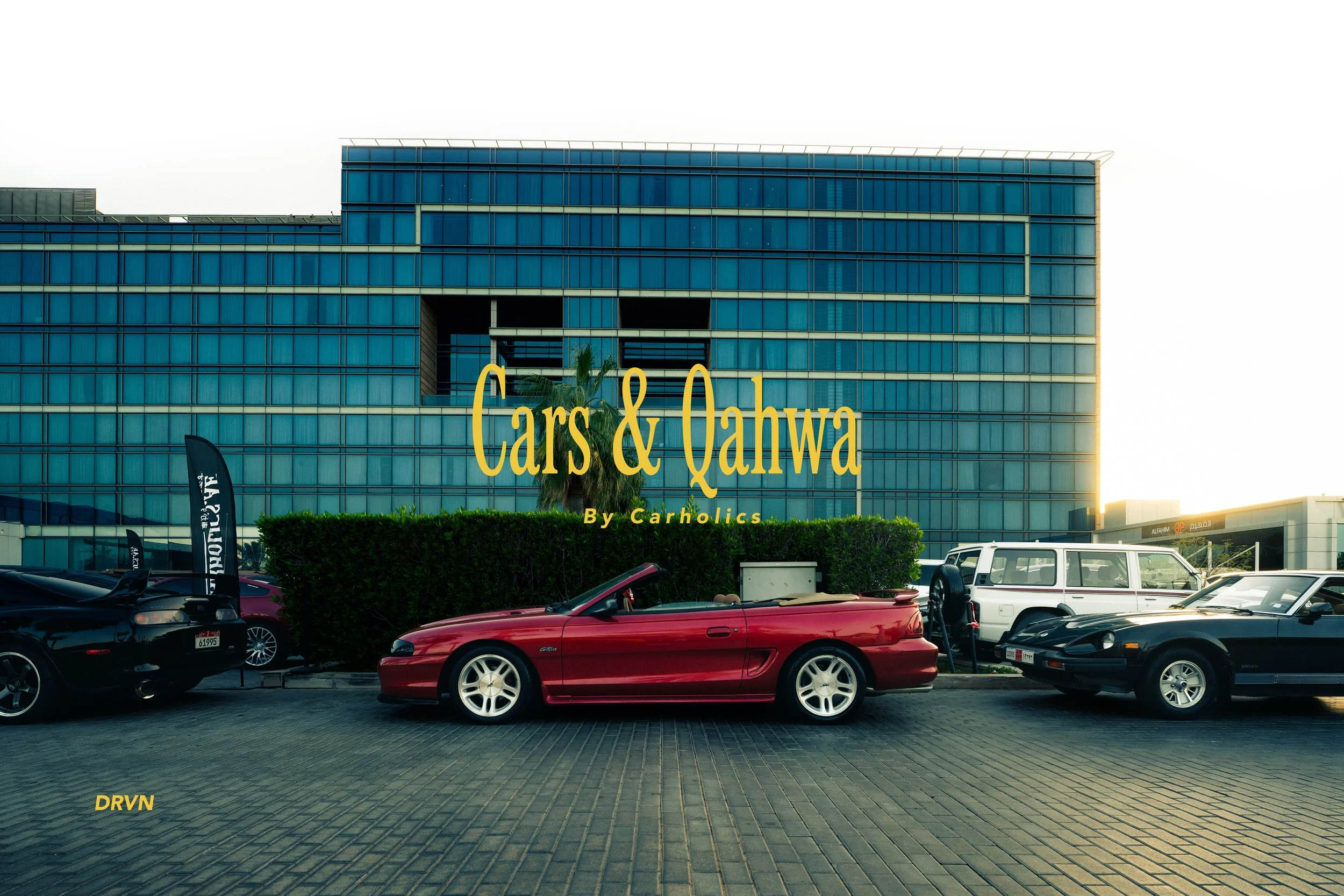 The image shows a modern glass building with the large text 'Cars & Qalwa' overlaid. In front of the building, there are several parked cars, including a red convertible, and some flags on the left side. A talk bubble with the word 'DRVN' is visible in the bottom left corner.