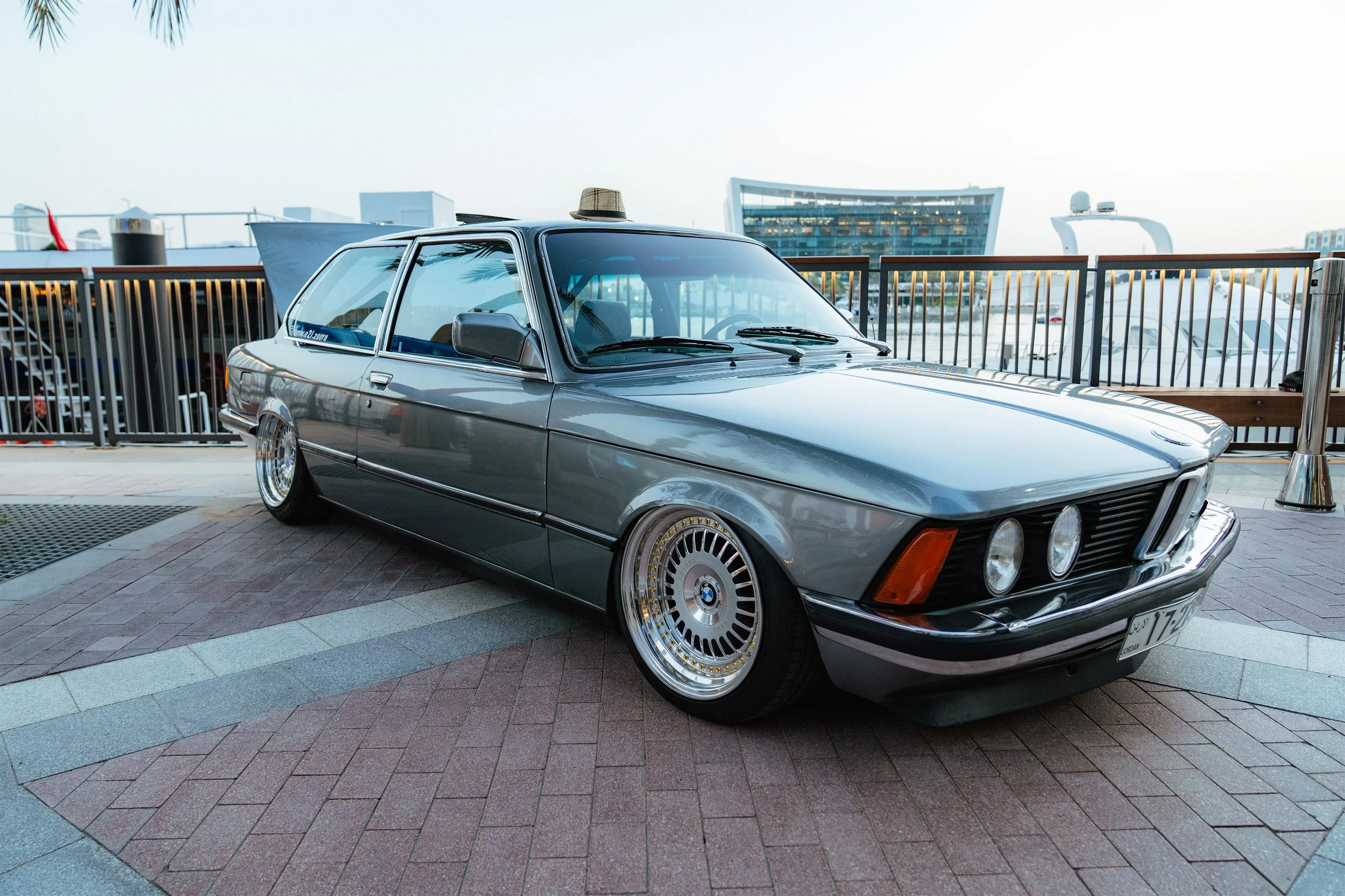 A vintage silver BMW car parked outdoors with a fedora hat placed on the roof. In the background, there are modern buildings and a railing, indicating an urban setting.