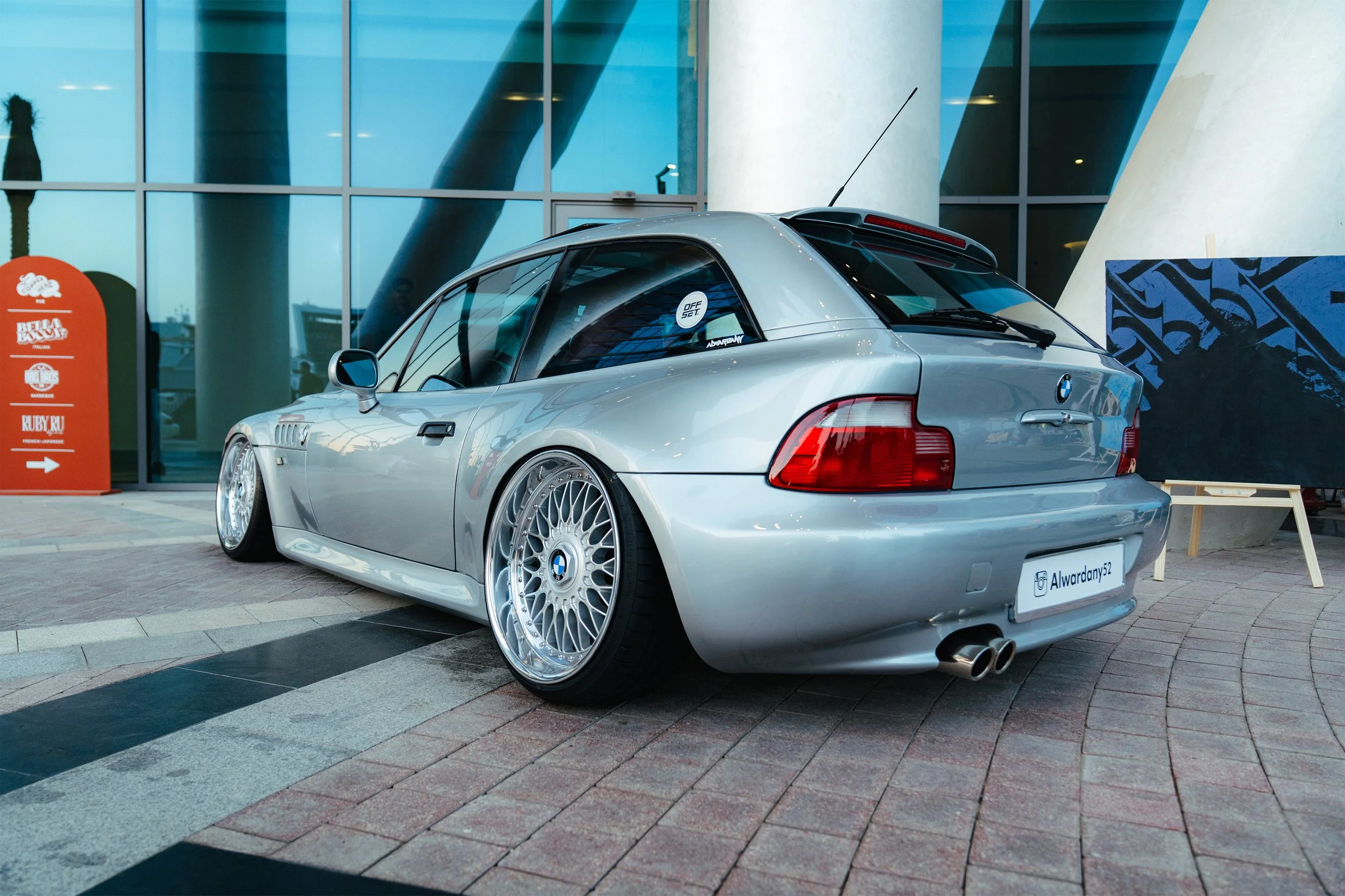 Silver BMW car with custom wheels parked outside a modern building with large glass windows and concrete pillars.
