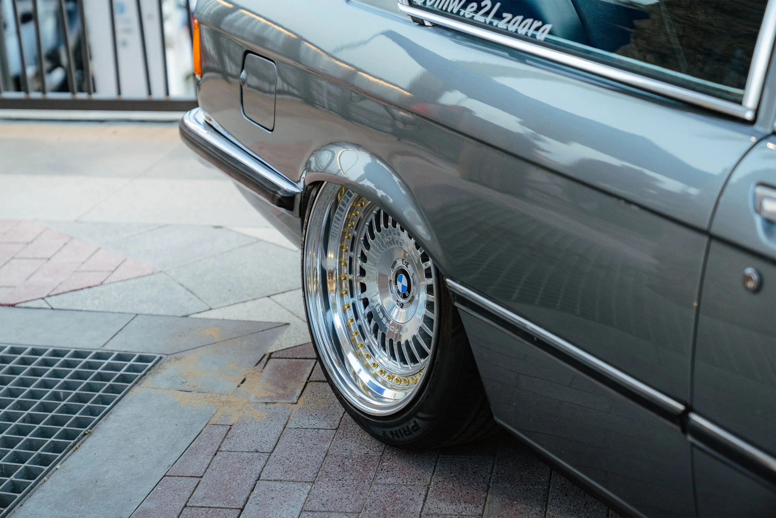 Close-up of the rear side of a grey BMW car, focusing on the wheel and part of the rear bumper, parked on a stone-paved surface.