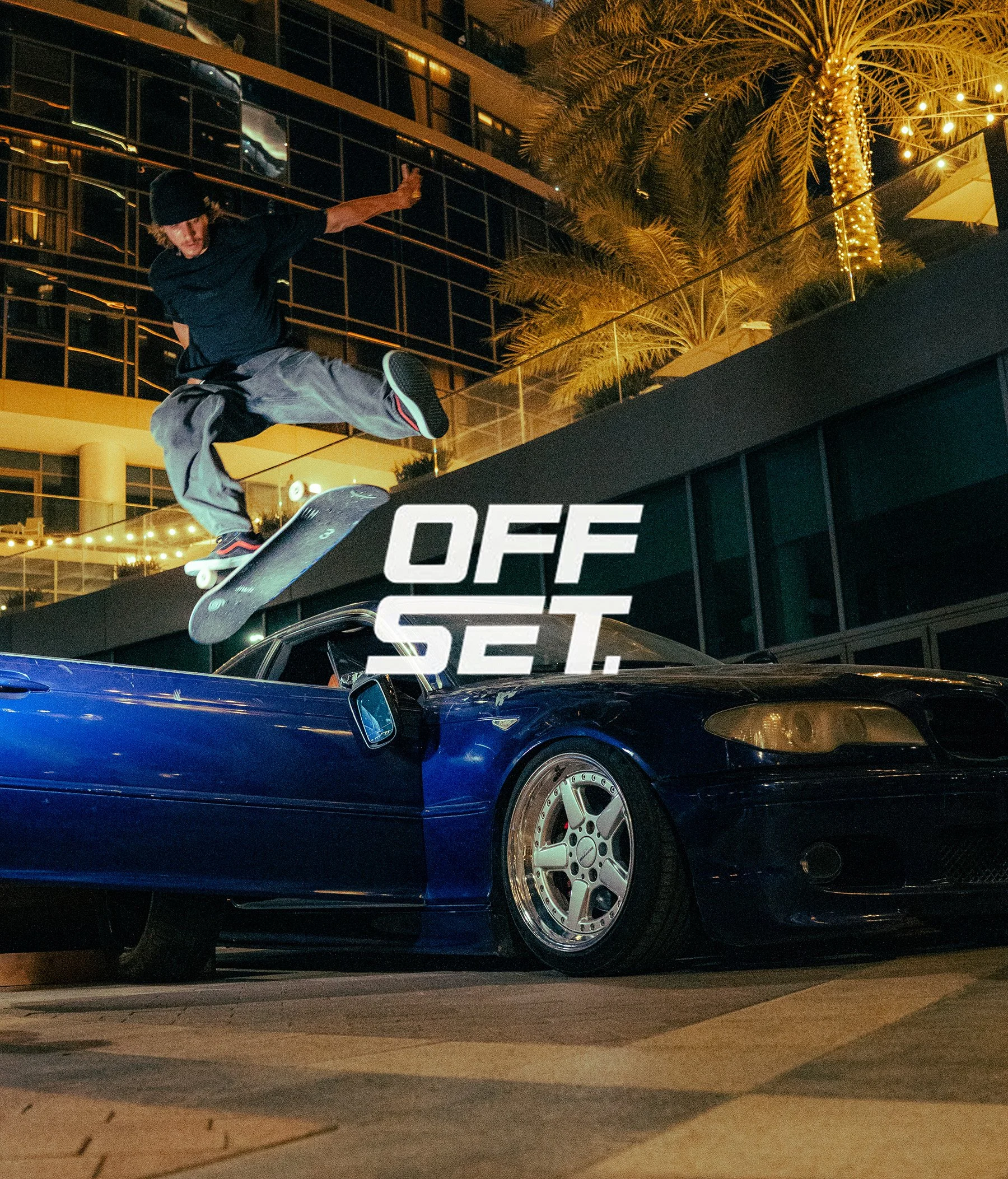 Skateboarder performing a trick at night in an urban setting with palm trees and building lights in the background.