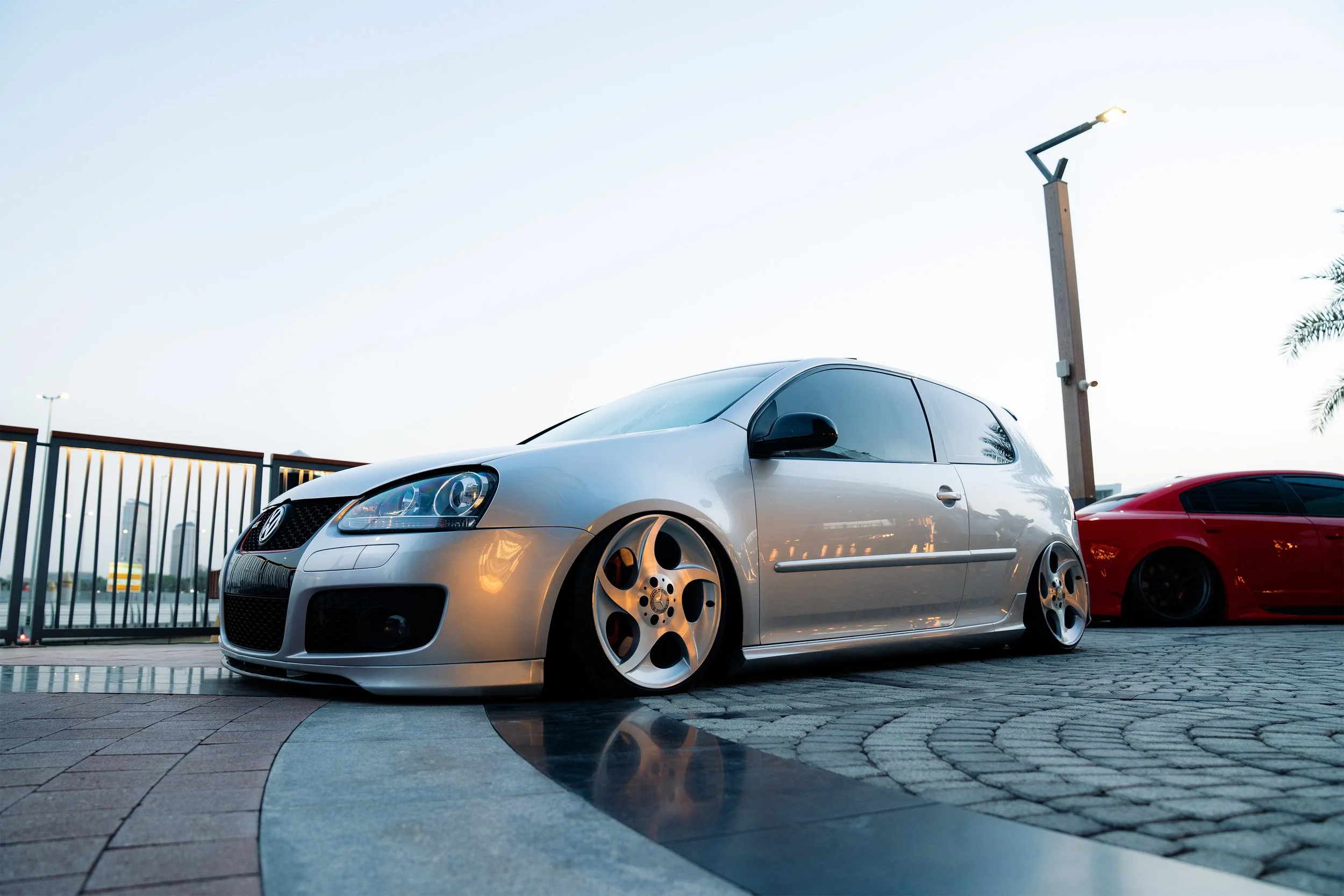 A silver sports car parked on a paved lot next to a red sports car with a fence in the background and a tall streetlight, under a clear blue sky.