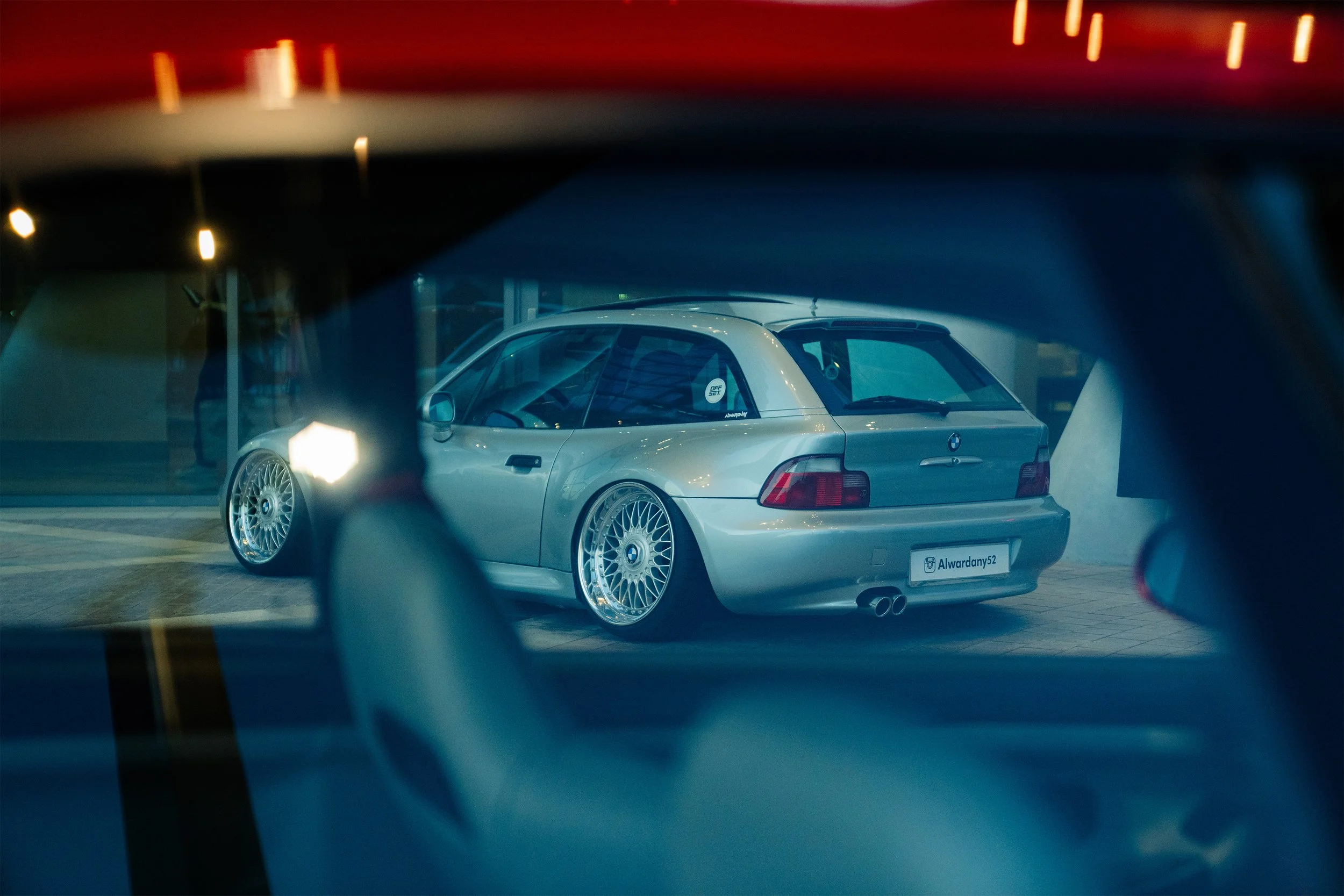 A silver BMW car with custom wheels and a spoiler, parked indoors, seen through the window of another vehicle