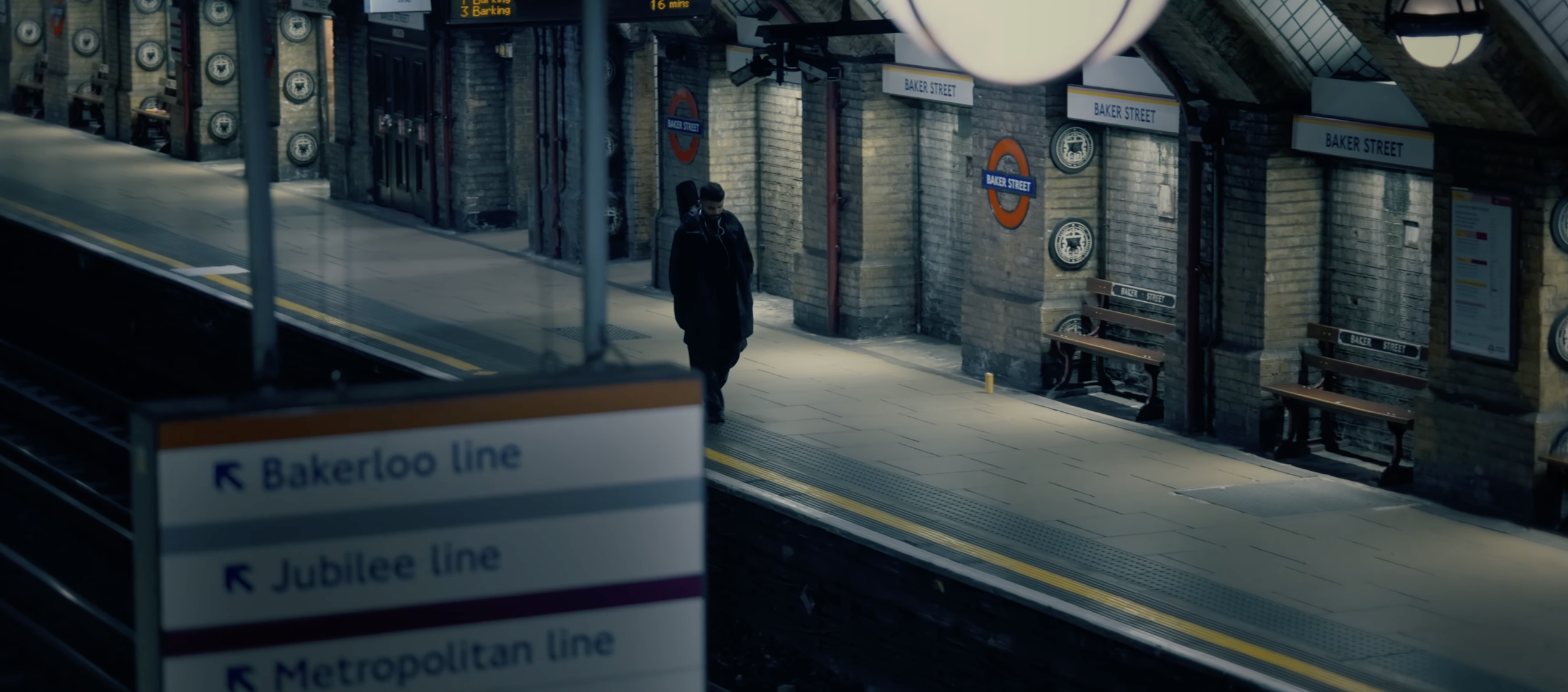 A person walking on a subway platform at Baker Street station in London, with brick walls, benches, and signs for the Bakerloo, Jubilee, and Metropolitan lines. The scene is mostly deserted with soft lighting.