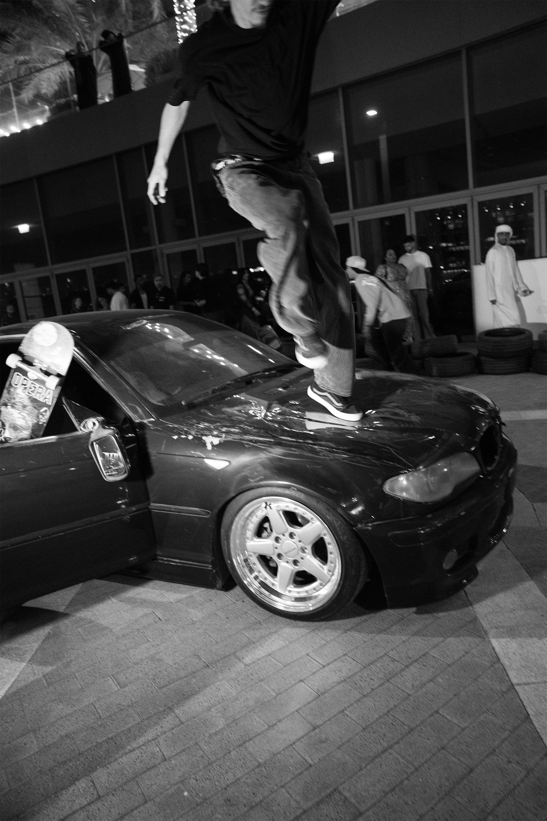 A person balancing on the hood of a black sports car at night, with a skateboard nearby and a crowd of people in the background.