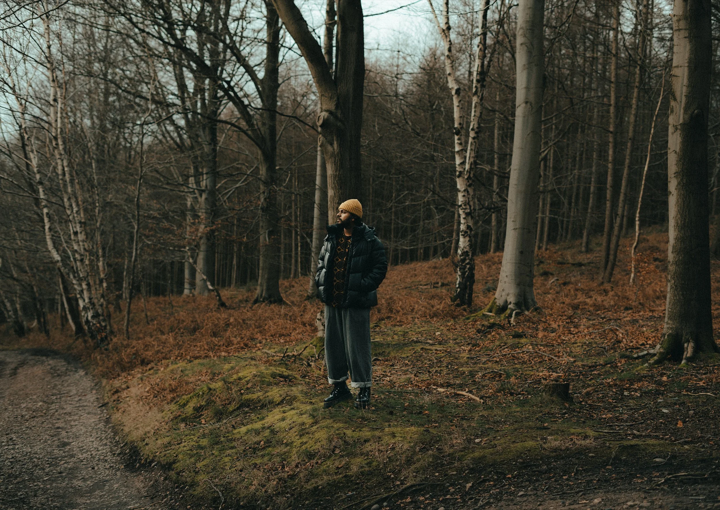 A man standing in a forested area with leafless trees, wearing a yellow beanie, glasses, a dark puffer jacket, and gray pants.
