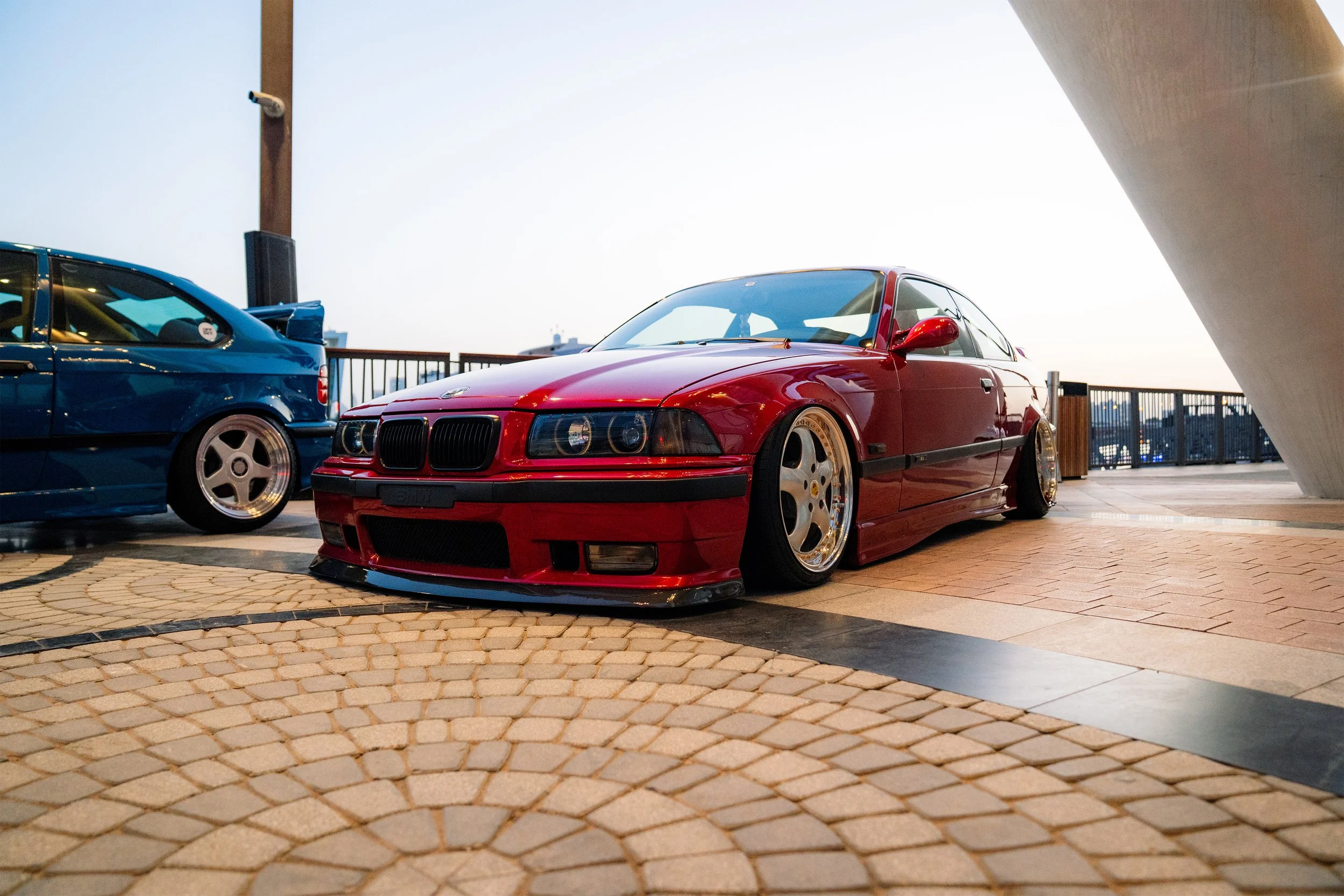 Red modified BMW car with lowered stance parked on cobblestone surface at sunset, next to a blue car, with a cityscape in the background.