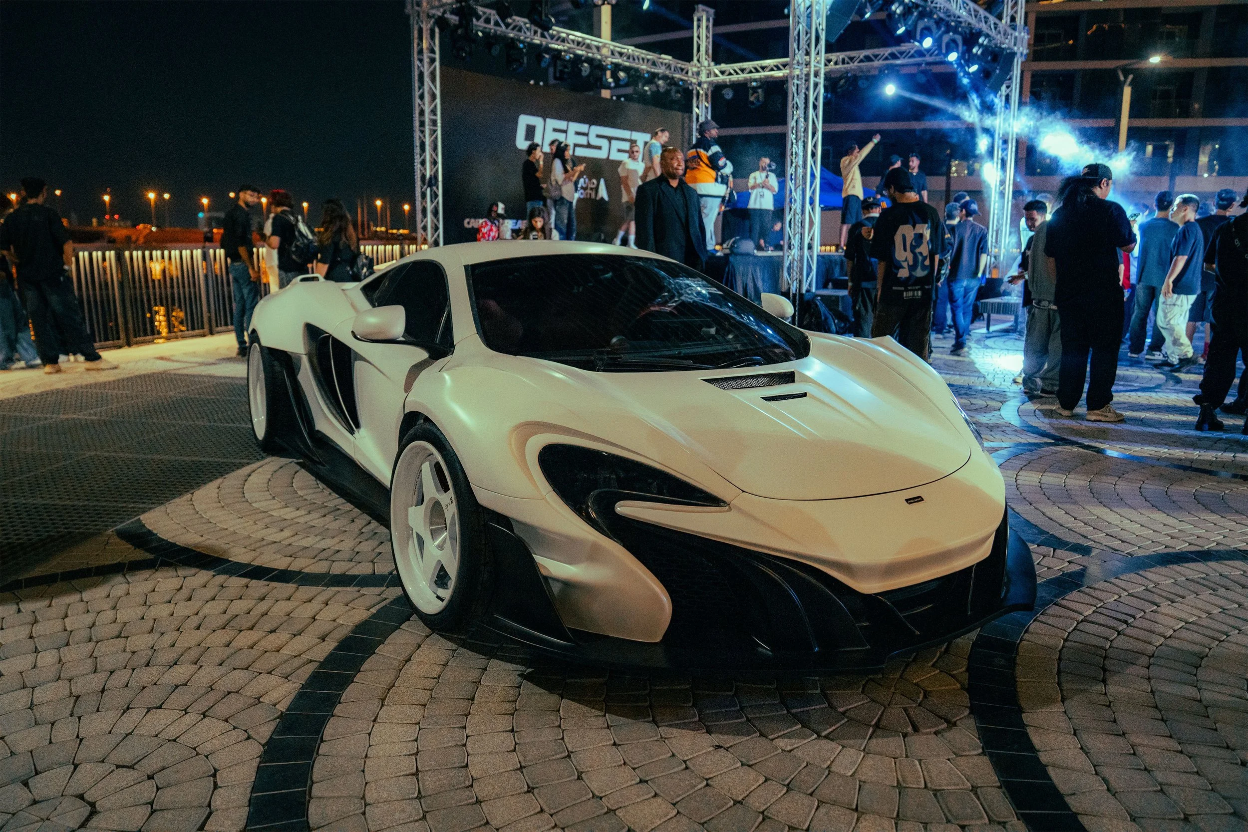 A white McLaren sports car parked in front of a crowd at a nighttime outdoor event with a stage and lights.