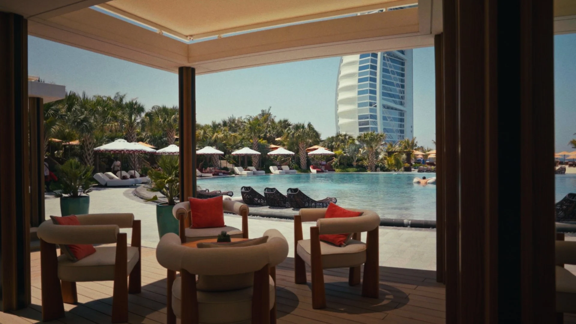 Indoor seating area with armchairs and cushions looking out to a pool with lounge chairs and umbrellas, palm trees, and a tall hotel building in the background.
