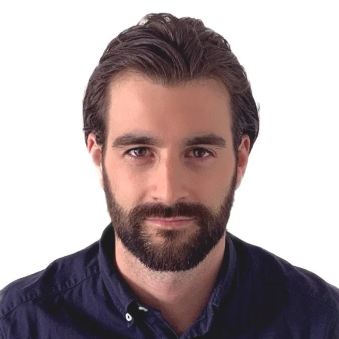 Portrait of a young man with dark hair and beard wearing a dark blue shirt, looking directly at the camera, against a white background.