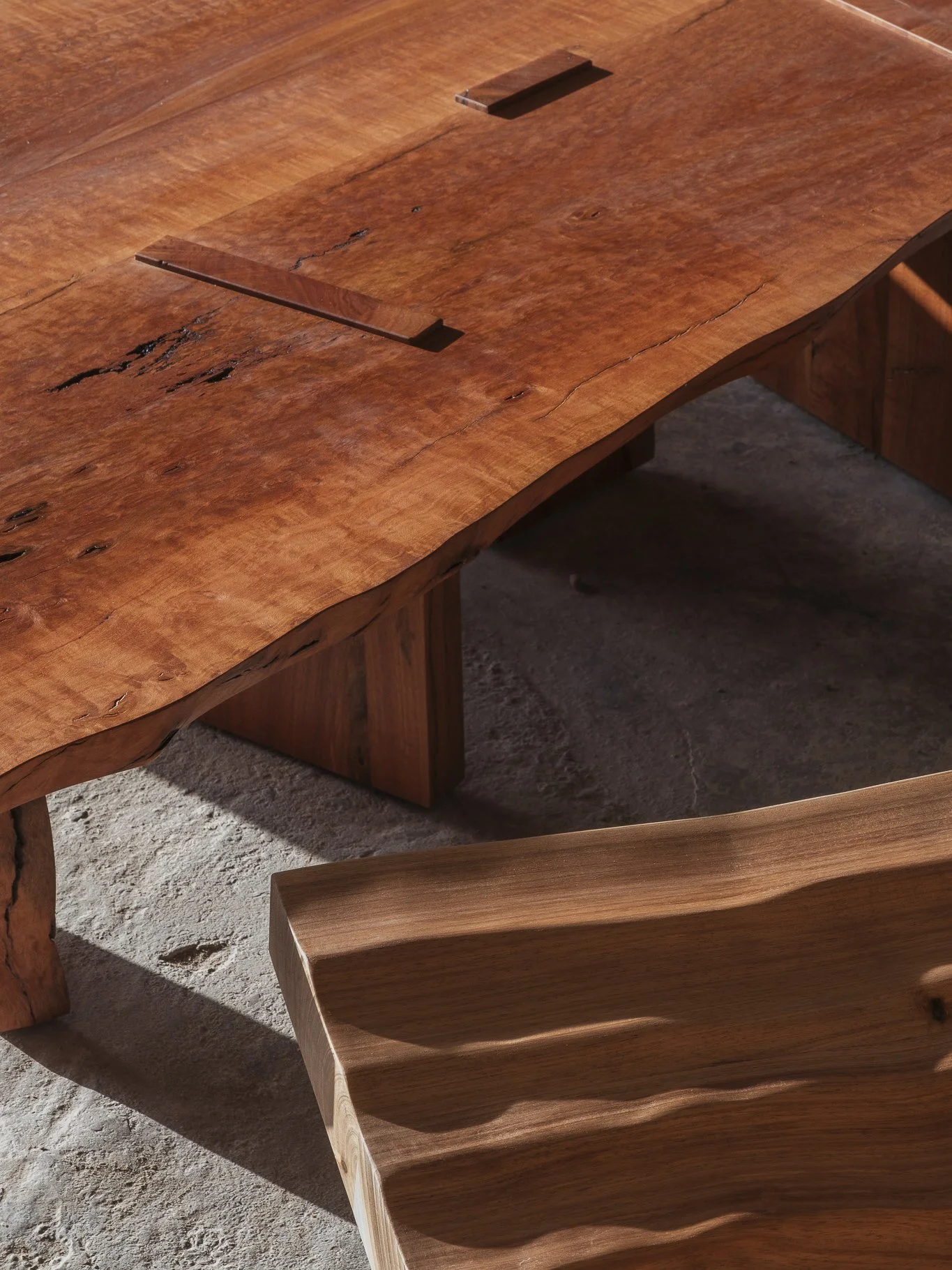 Mesa de madeira com acabamento rústico, com um tampo de madeira natural com imperfeições e uma estrutura de suportes também de madeira. Ao lado, uma cadeira de madeira com acabamento mais suave e uma superfície com várias reentrâncias onduladas.