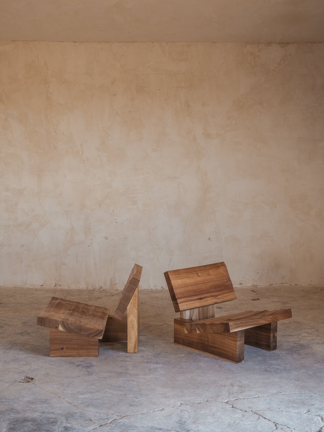 Duas cadeiras de madeira com design minimalista e uma textura arrojada, em cima de um piso de cimento, contra uma parede de cor bege clara.