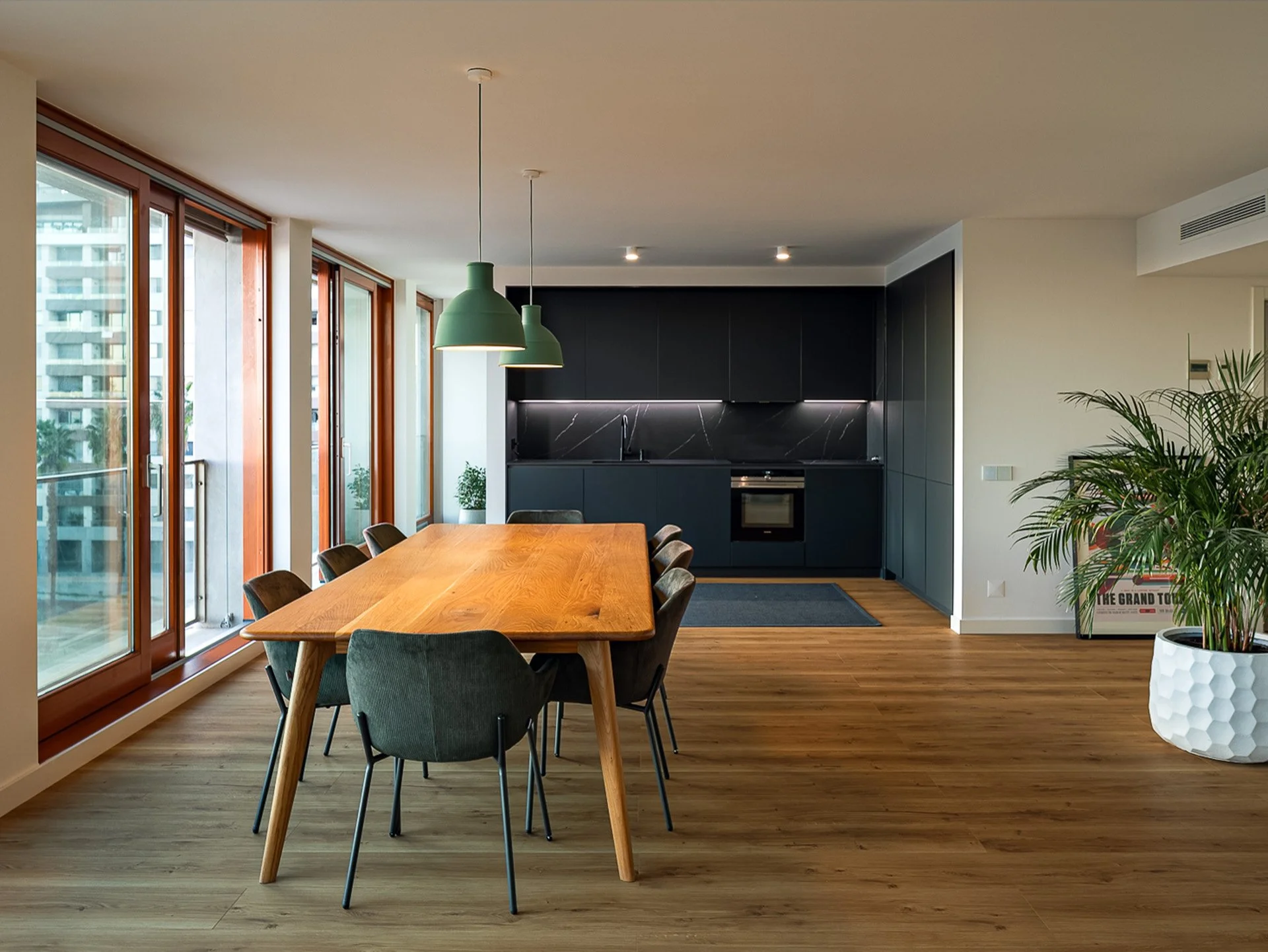 Sala de jantar moderna com mesa de madeira maciça e oito cadeiras de tecido verde escuro, cozinha ao fundo com armários pretos e bancada de mármore preto, grandes janelas de vidro com molduras de madeira, piso de madeira clara e uma grande planta.