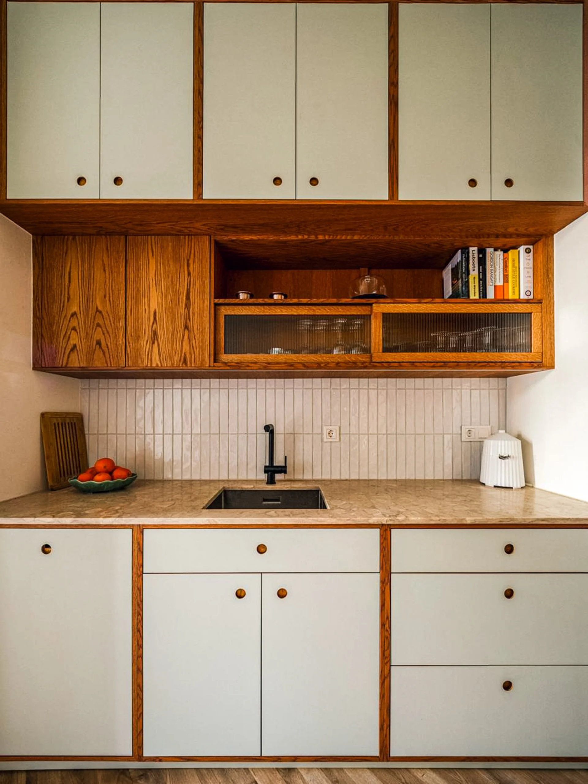 Cozinha feita à medida com armários de madeira e portas brancas, lava louças preto, balcão de mármore com tigela de laranjas, utensílios de madeira, livros na prateleira superior e um aparelho de cozinha no canto direito.