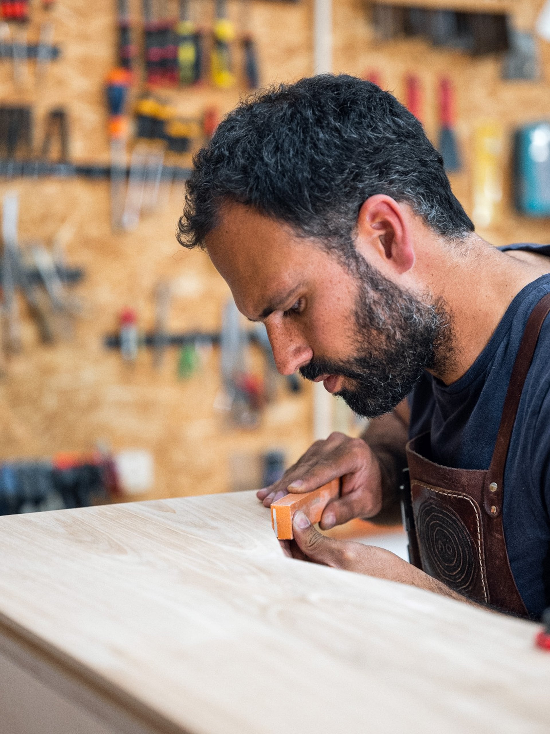 Zé a lixar uma superfície de madeira no atelier Nó, com ferramentas ao fundo.